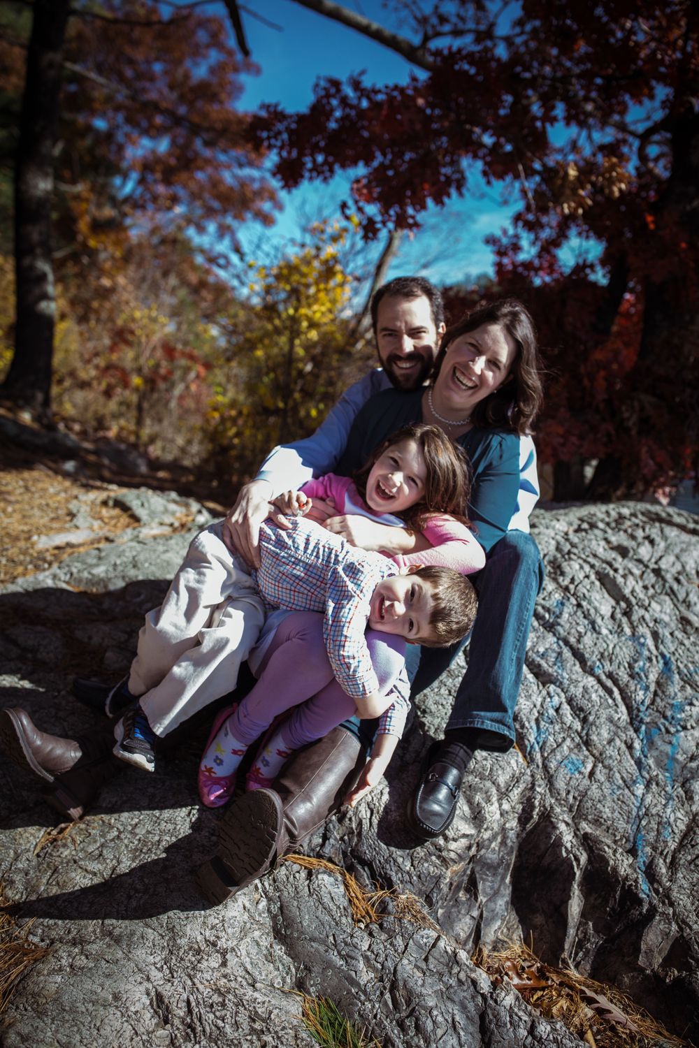 a family of four, sitting on a rock with autumn leaves on the trees behind them. all are hugging