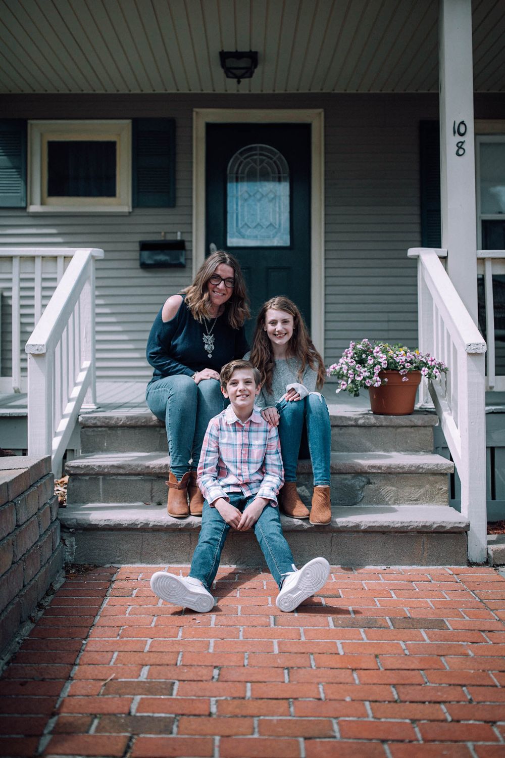 a family of three, a mom, a daughter and a son, sitting on their front porch steps together, smiling