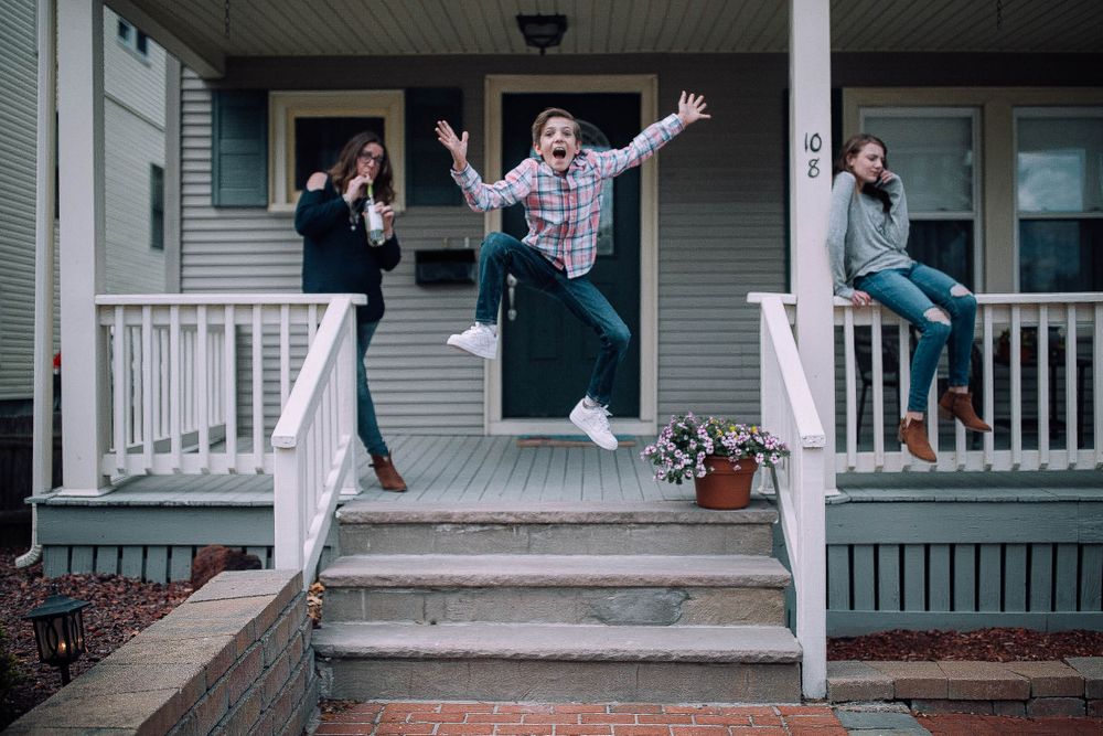 a family of three, a mom with wine, a daughter on her phone, and a son leaping off their front porch, acting silly