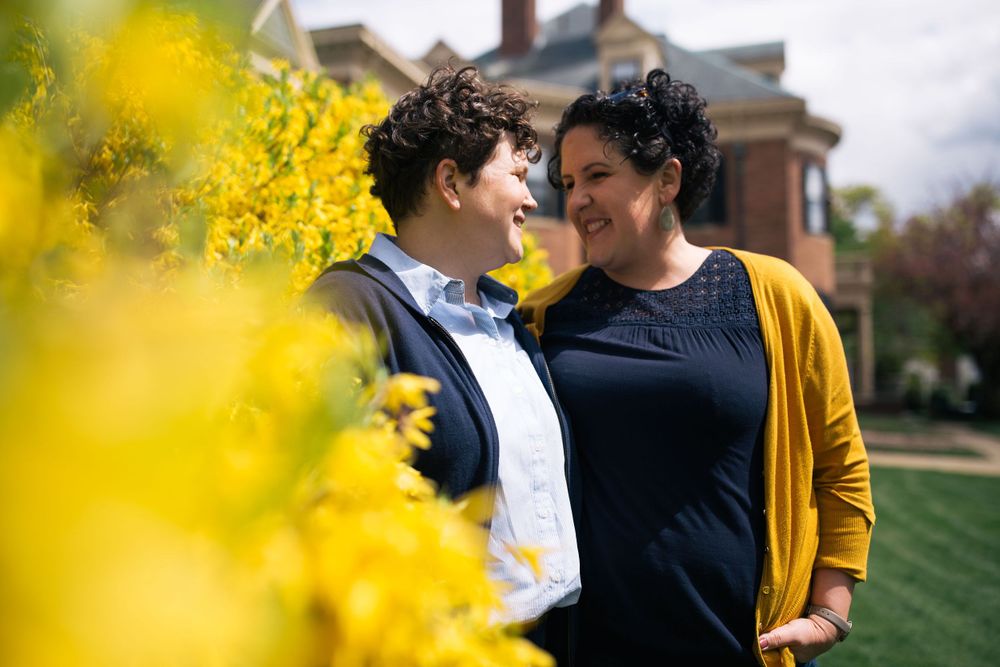 a queer couple hold each other in front of a yellow forsythia bush