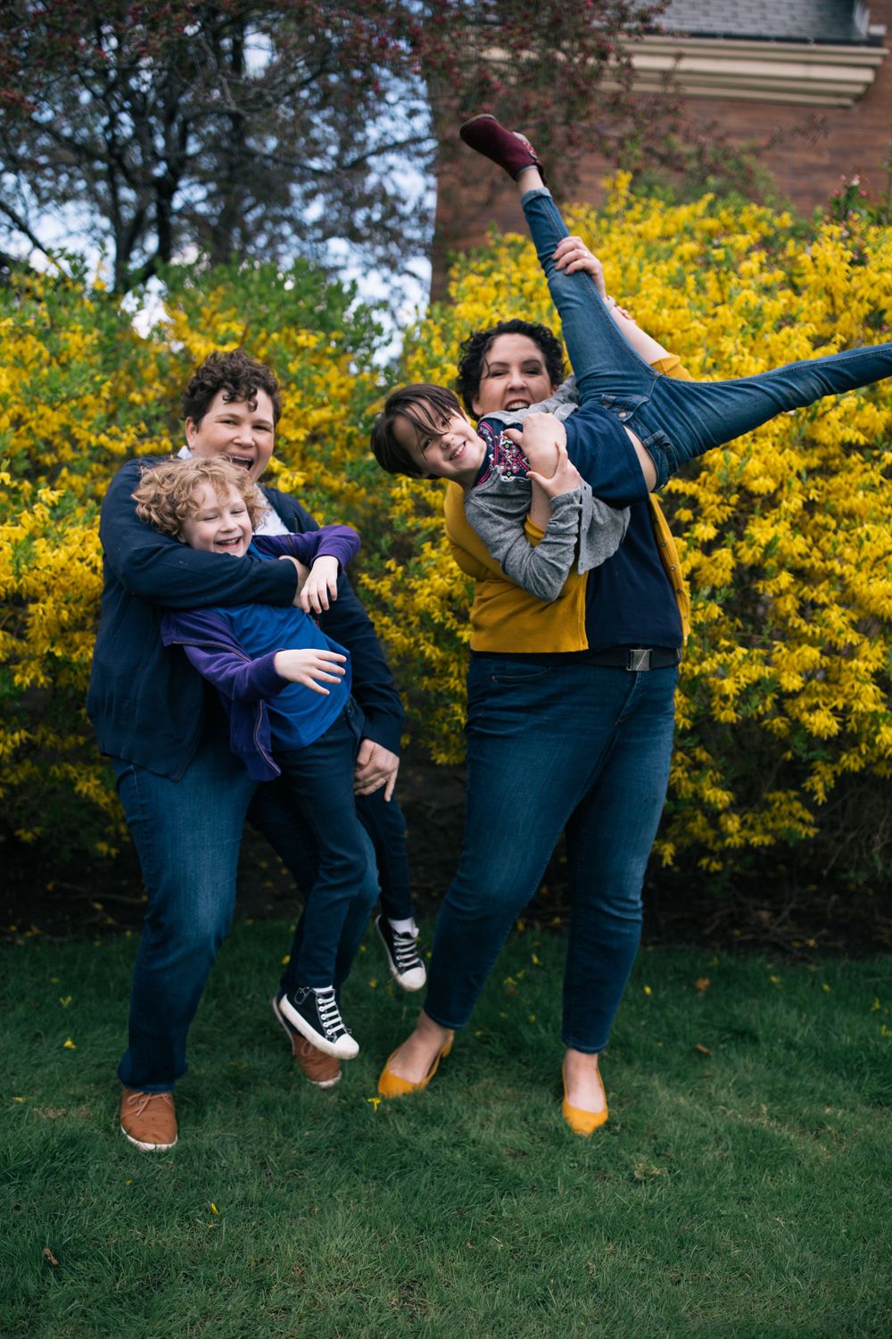 two moms holding their kids upside down and acting silly in front of a forsythia bush with green grass