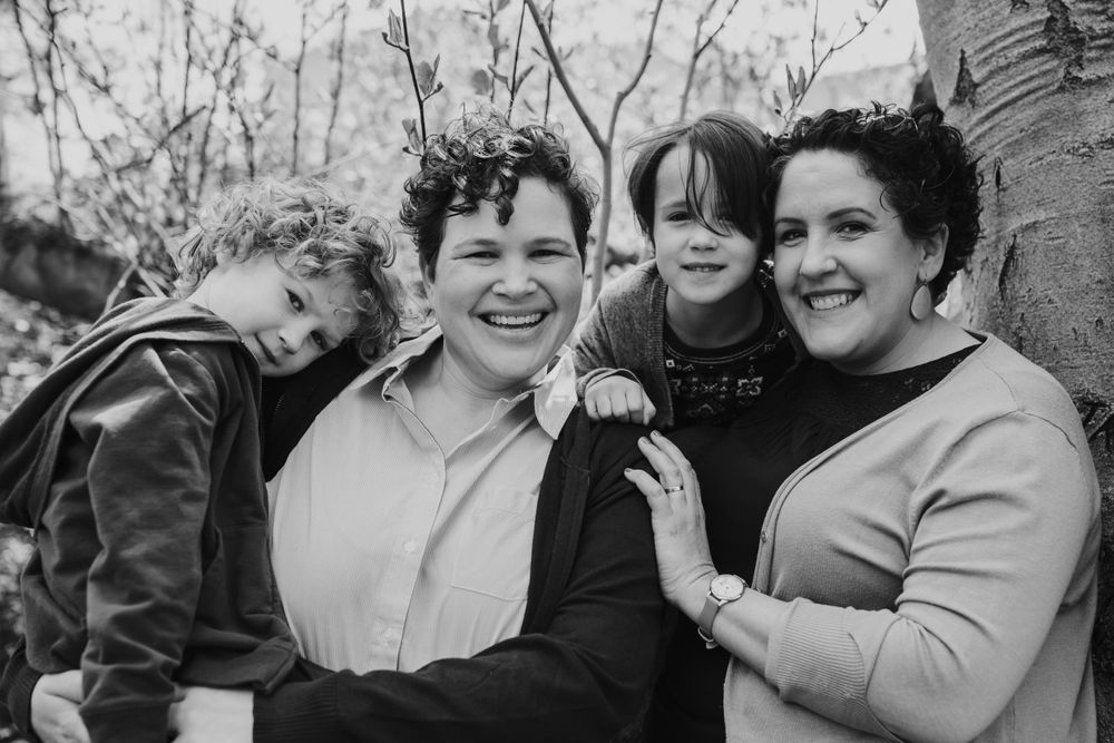 black and white image of a queer family, with two moms and two kids smiling widely under a tree