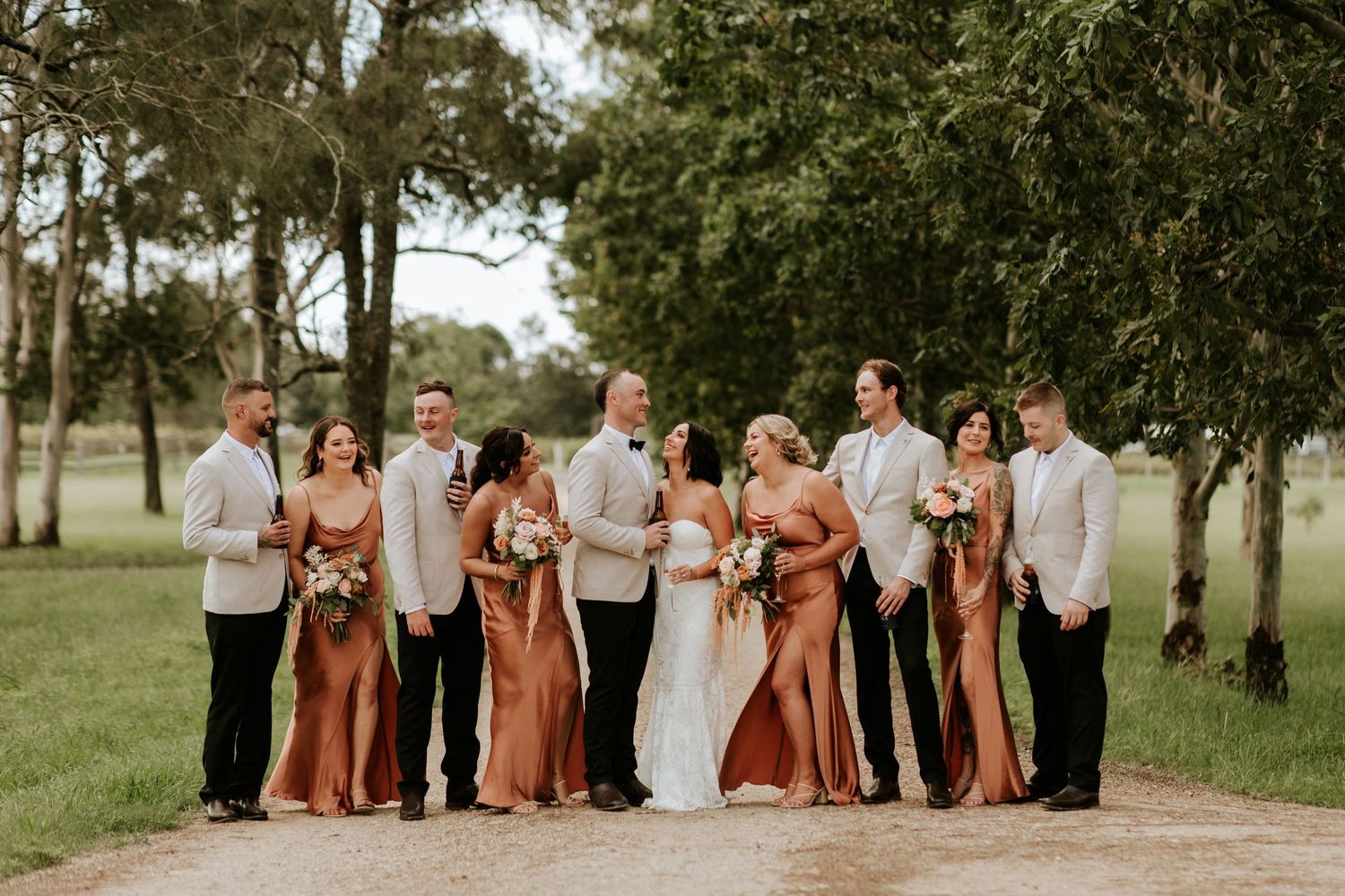 Wedding party poses on a path with bridesmaids in copper-colored dresses and groomsmen in light suits.