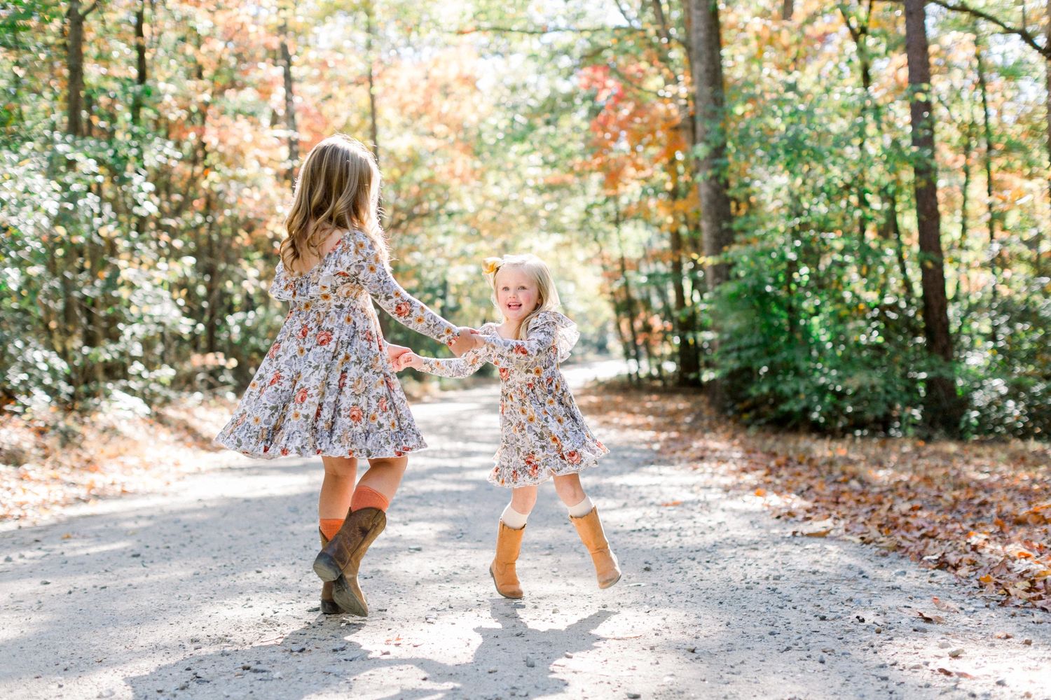Two young girls in matching floral dresses dancing on a sunlit autumn forest path.