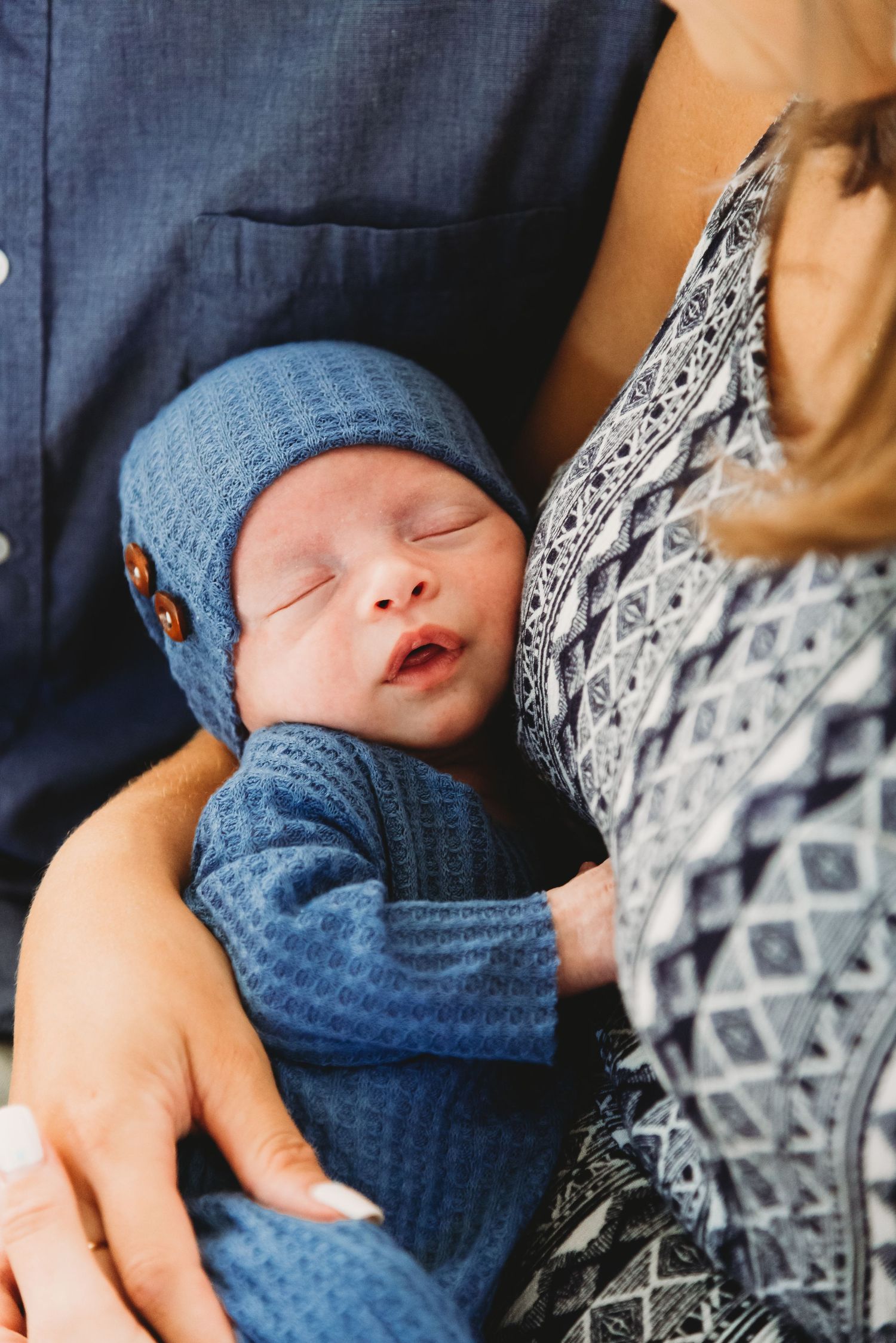Sleeping baby in blue knit outfit and hat rests peacefully against parent.