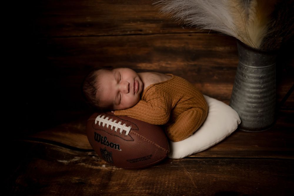 A sleeping newborn rests on a football against a dark wooden backdrop.