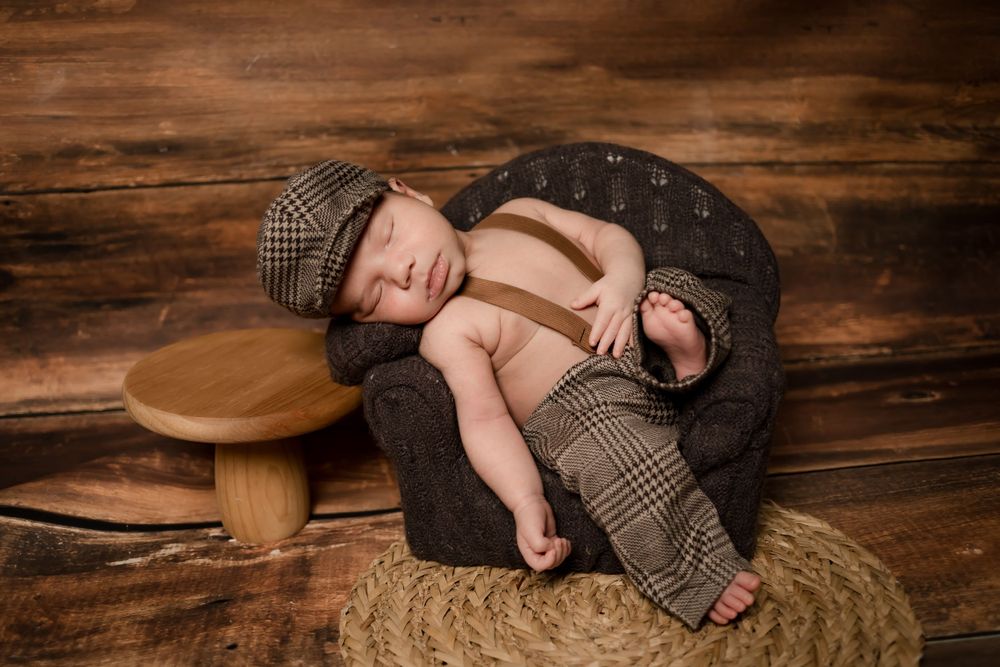 A sleeping newborn wearing vintage-style clothing and cap poses on textured fabric.