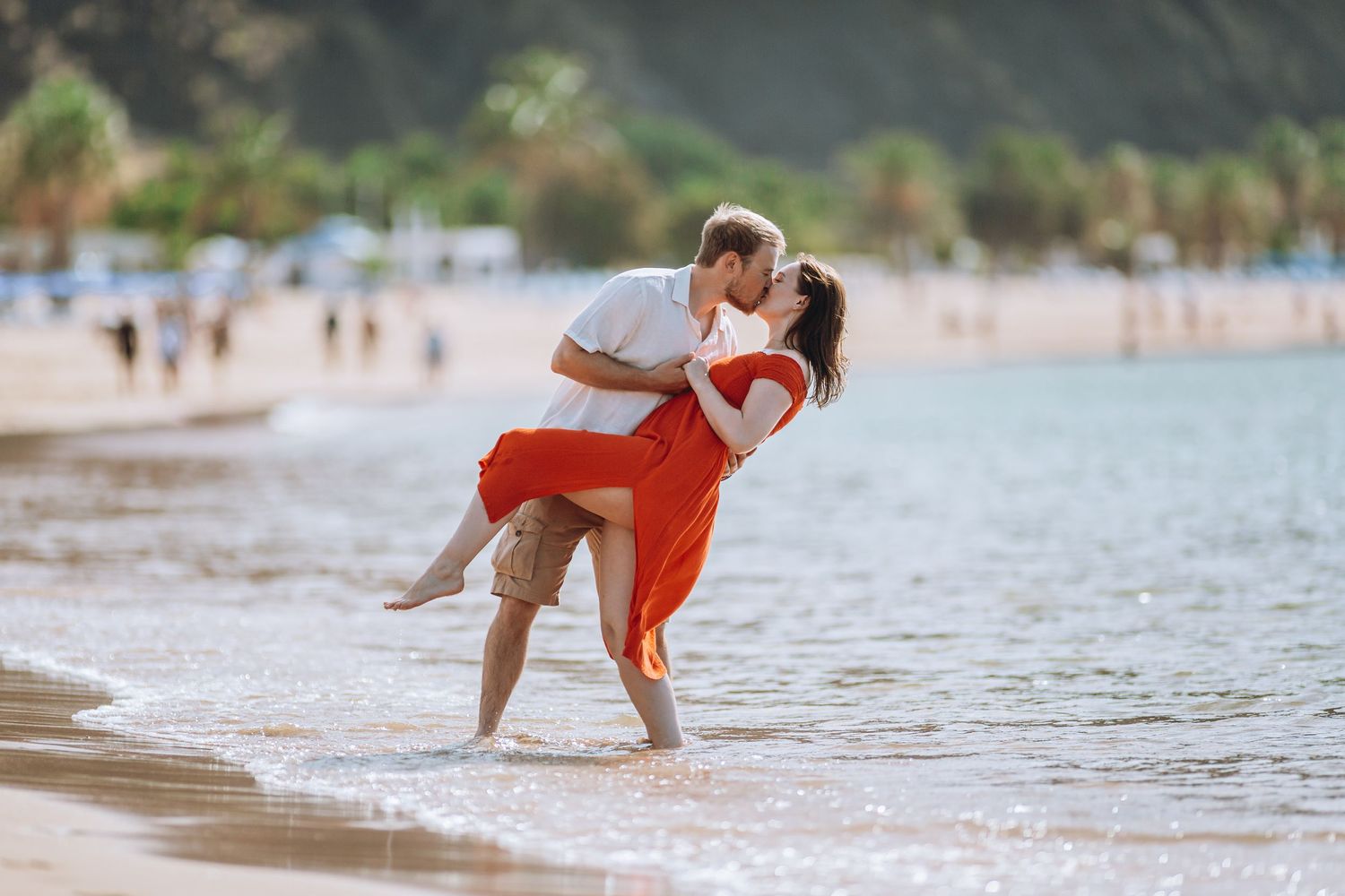 Playful couple dancing and twirling on sandy beach with ocean waves in background.