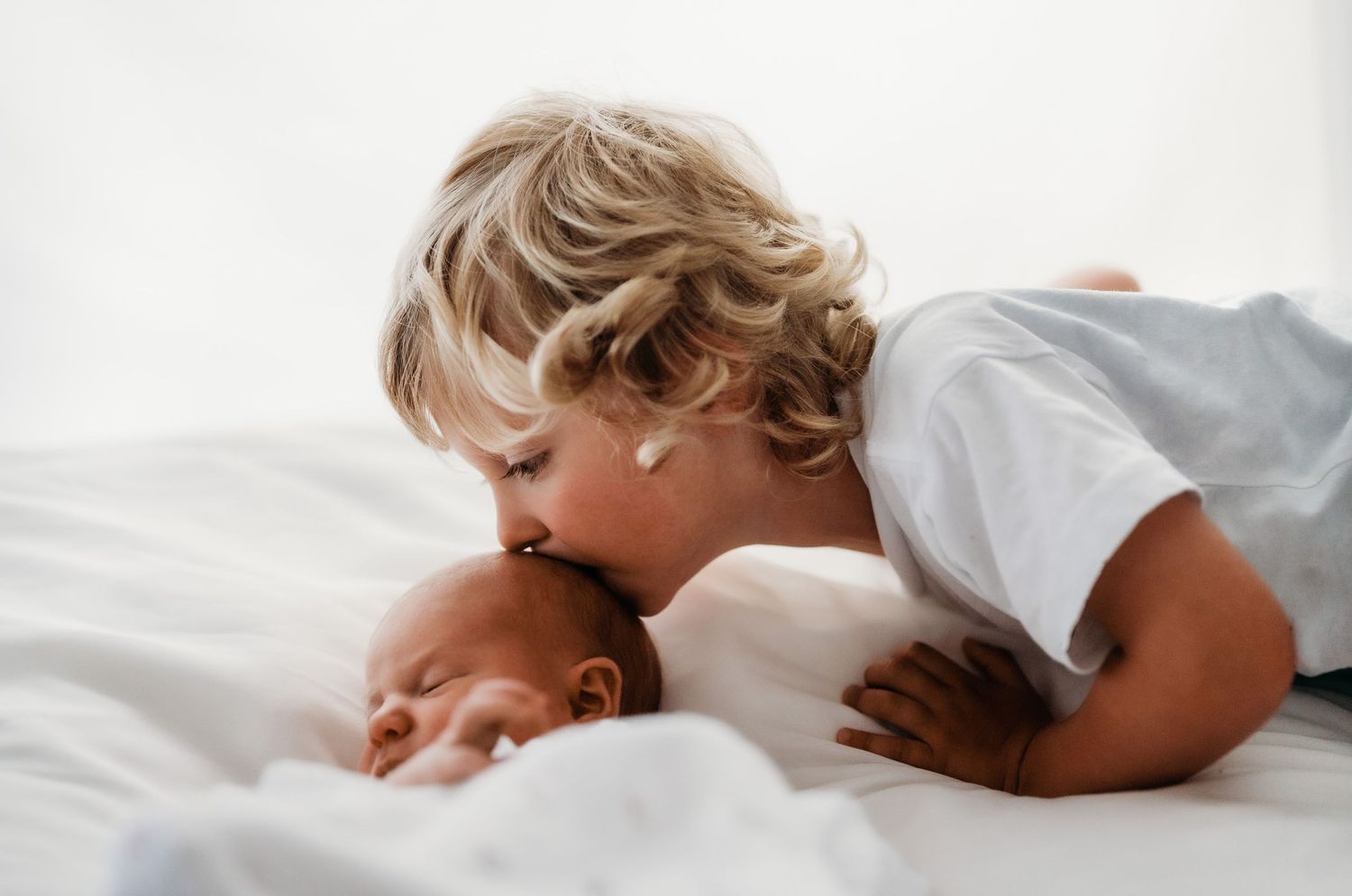 Classic white image of a newborn baby and her older brother, softly kissing her on the forehead.