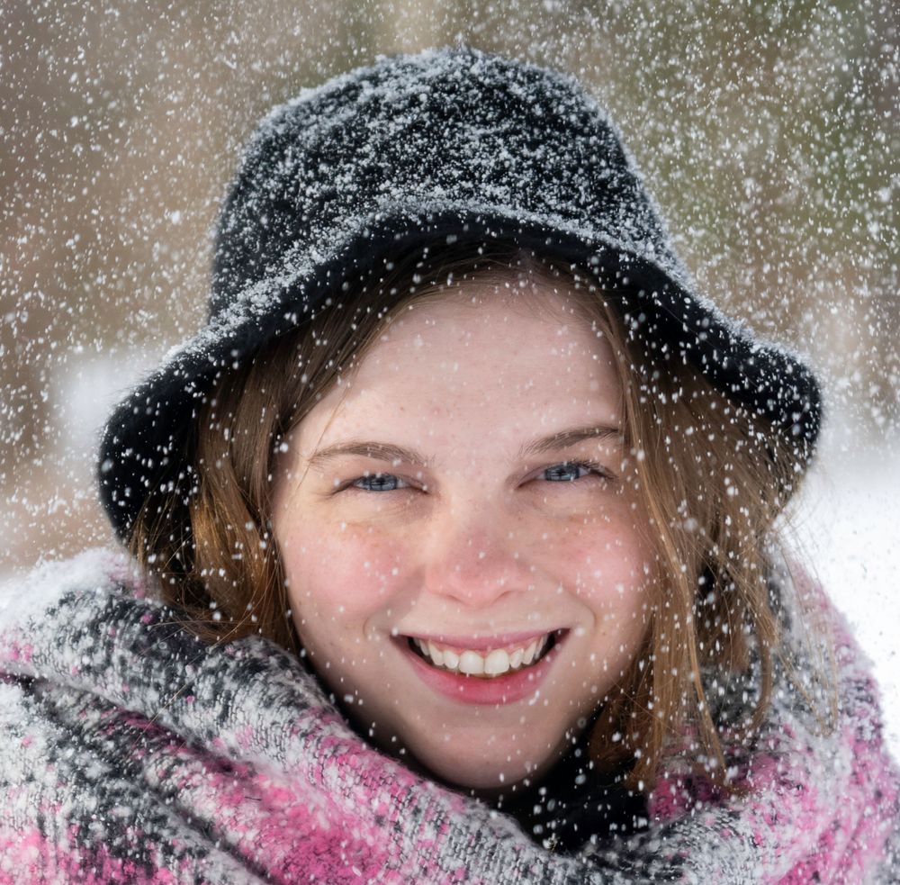 Person in a black winter hat smiling while snowflakes fall around them on a snowy day.