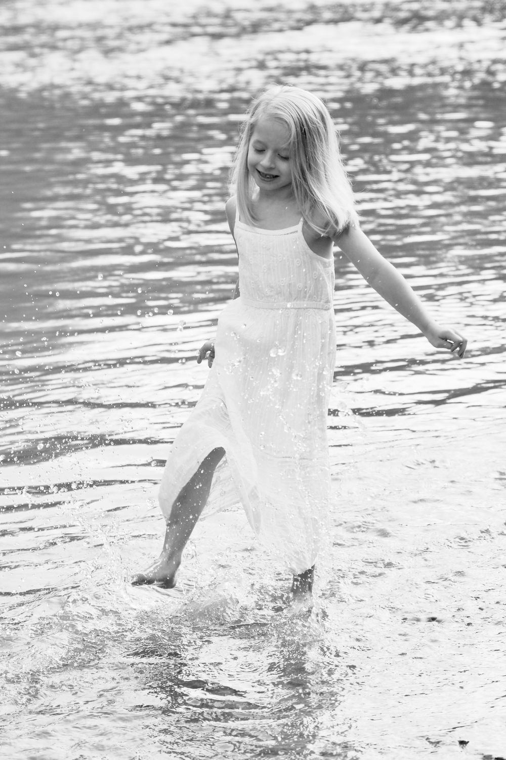 Black and white image of child splashing in shallow water wearing white dress.