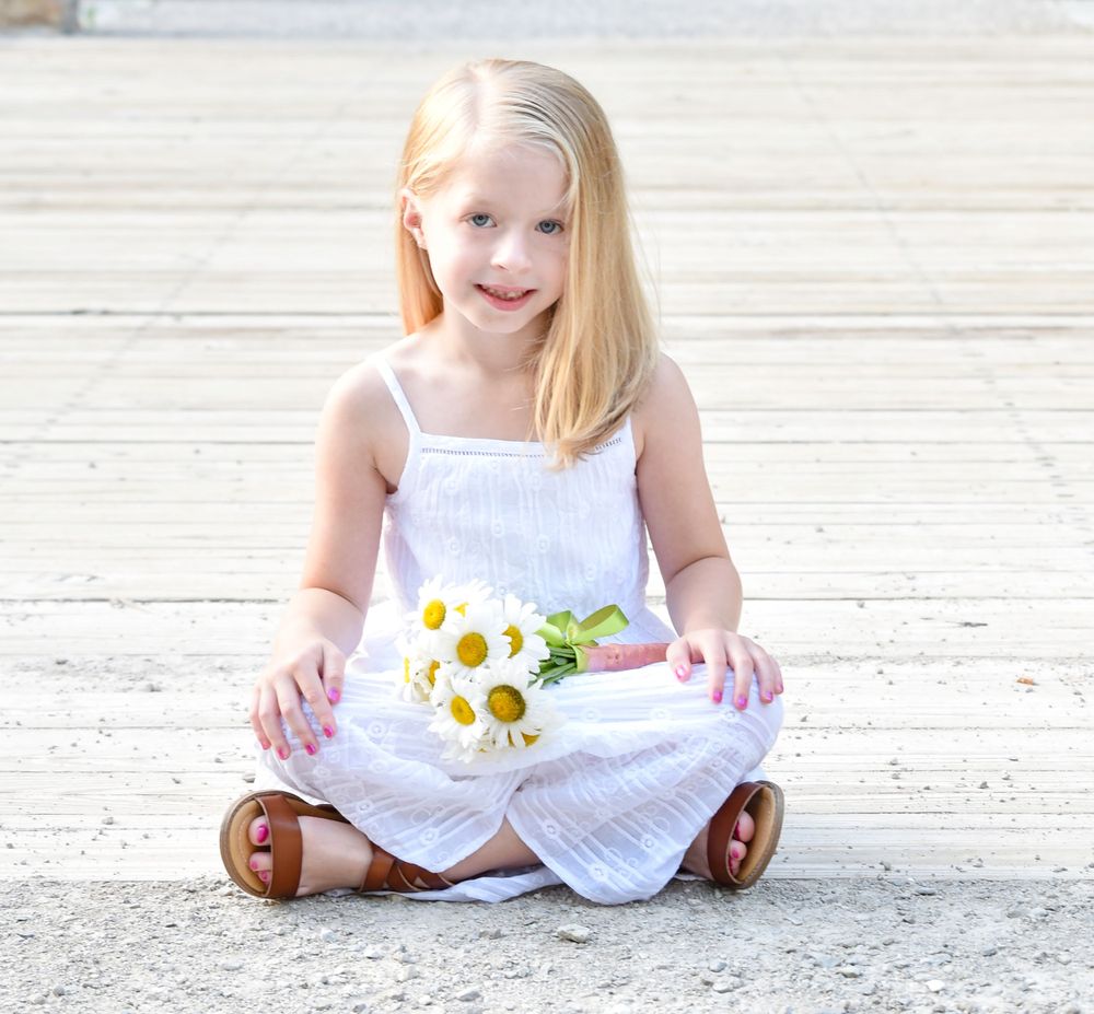 Young child in white dress sitting cross-legged holding daisies on wooden deck.