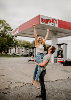 A couple strikes a joyful pose with lifted arms at a vintage Stop-N-Buy station.