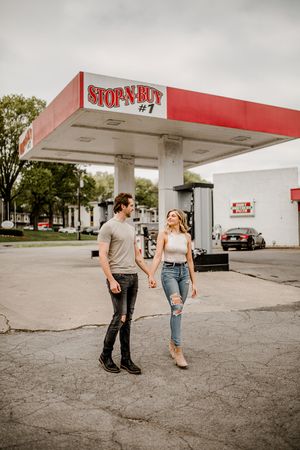 A couple holding hands walks together at a vintage Stop-N-Buy gas station on an overcast day.
