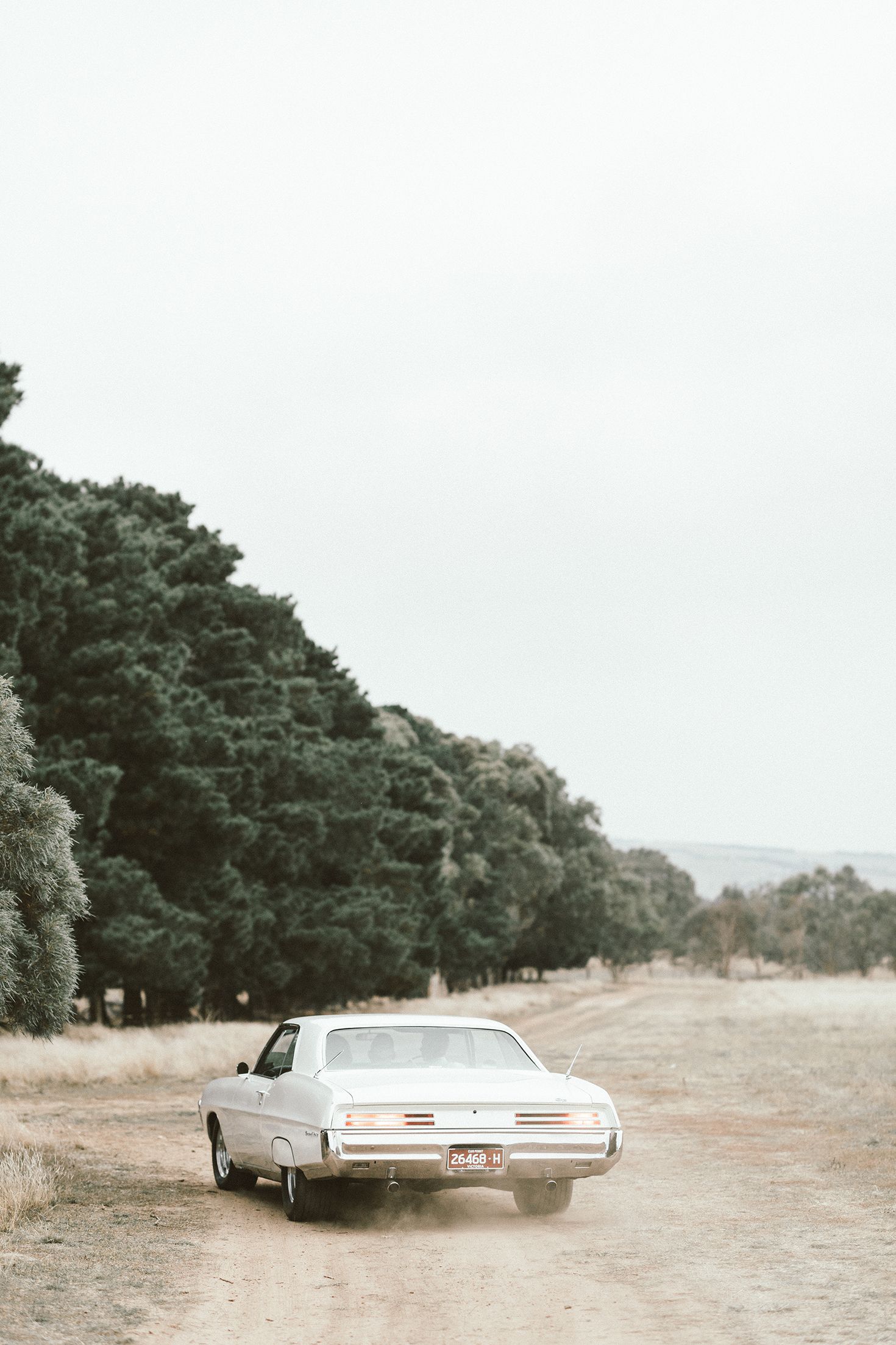 A classic white vintage car parked on a dirt road with trees in the background.