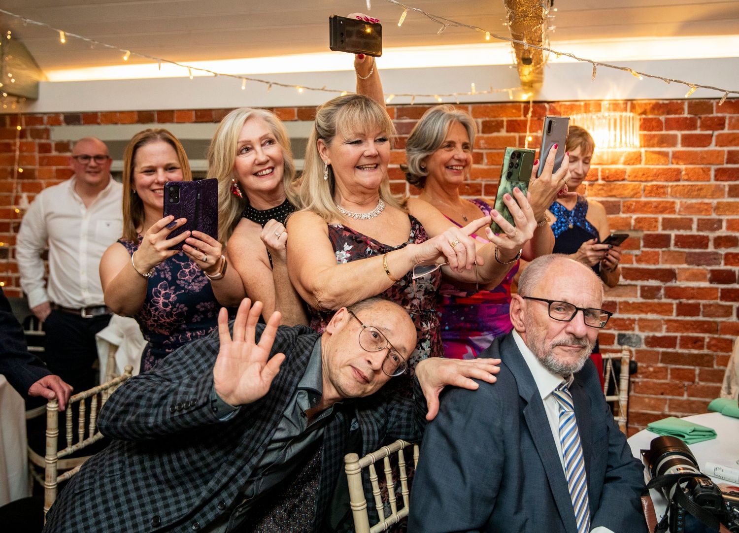 Wedding guests celebrating and laughing together at a reception with brick wall backdrop.