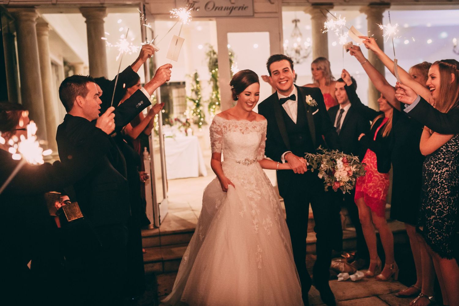 Bride and groom exit wedding venue through sparkler tunnel surrounded by celebrating guests at night.