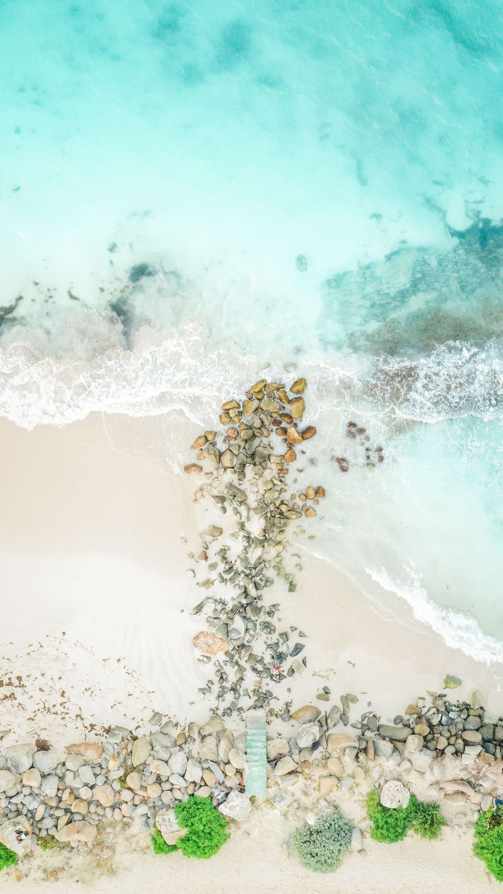 Aerial view of turquoise ocean waves meeting white sandy beach with scattered rocks and green vegetation.