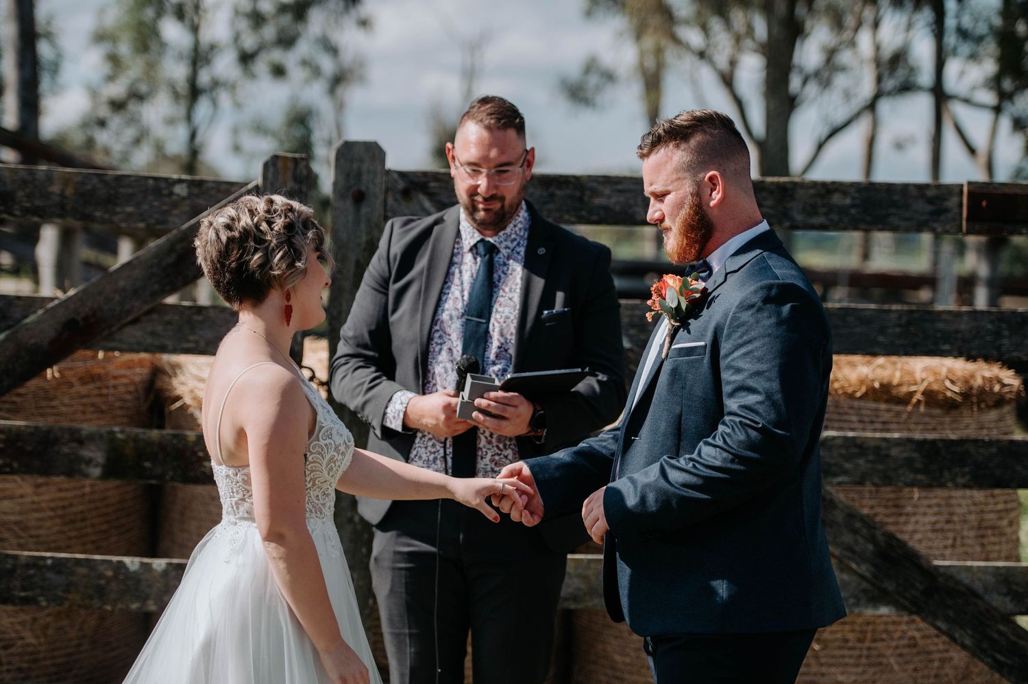 A wedding ceremony takes place in front of a rustic wooden fence with an officiant presiding.