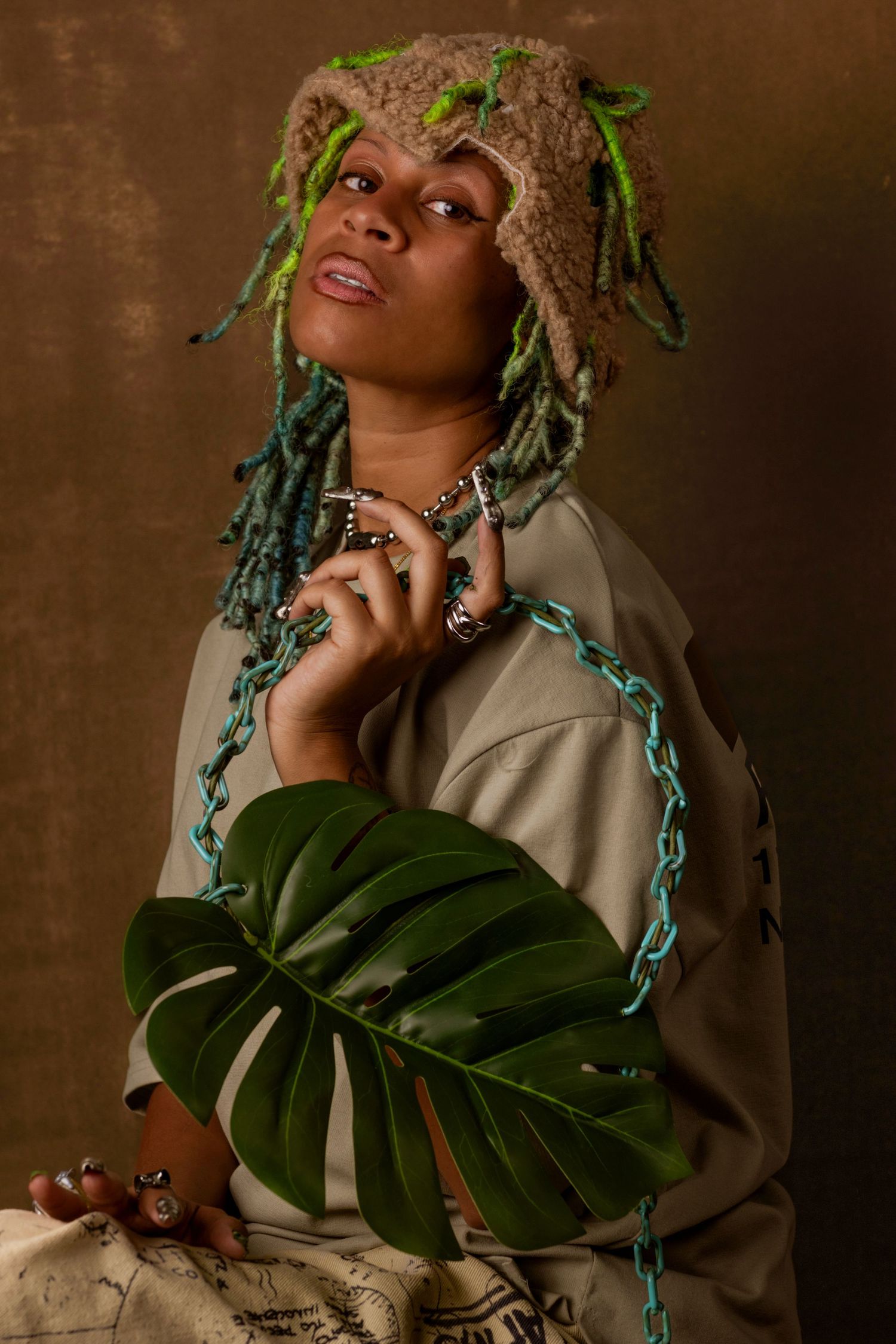 Artistic portrait featuring a person with green foliage accents holding a large monstera leaf against a brown backdrop.