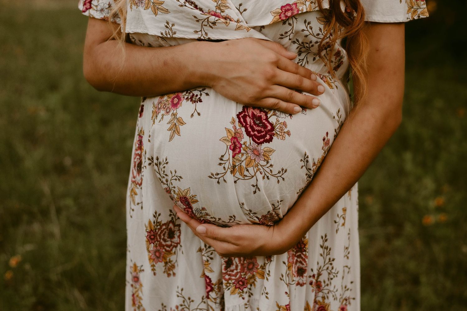 Close-up detail of baby bump in white floral maternity dress against natural outdoor backdrop.