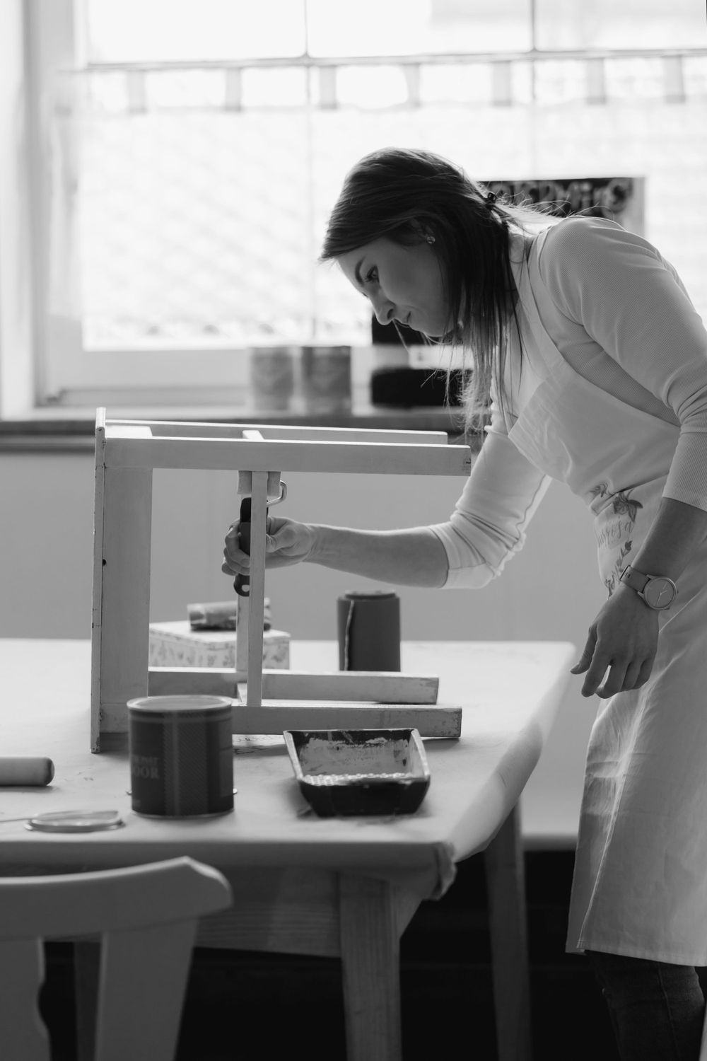Künstler im weißen Kittel arbeitet sorgfältig an einem Arbeitstisch mit Materialien und Werkzeugen in Schwarz-Weiß-Fotografie.