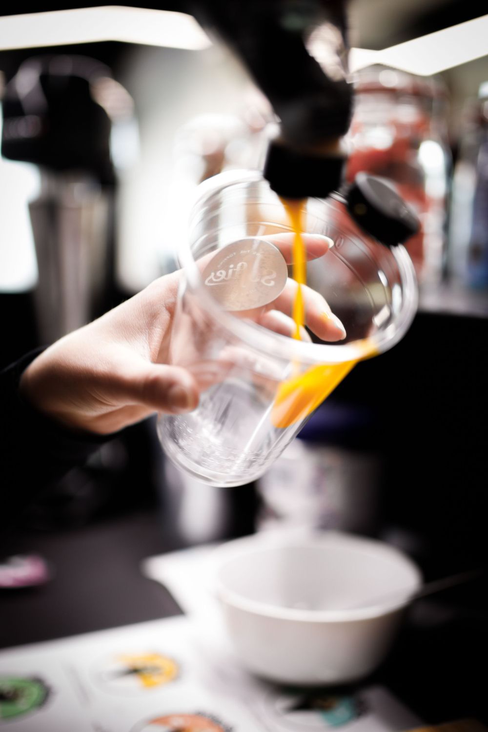 Eine verschwommene Bewegungsaufnahme von Händen, die in einer Bar orangefarbene Flüssigkeit in einen durchsichtigen Plastikbecher gießen.