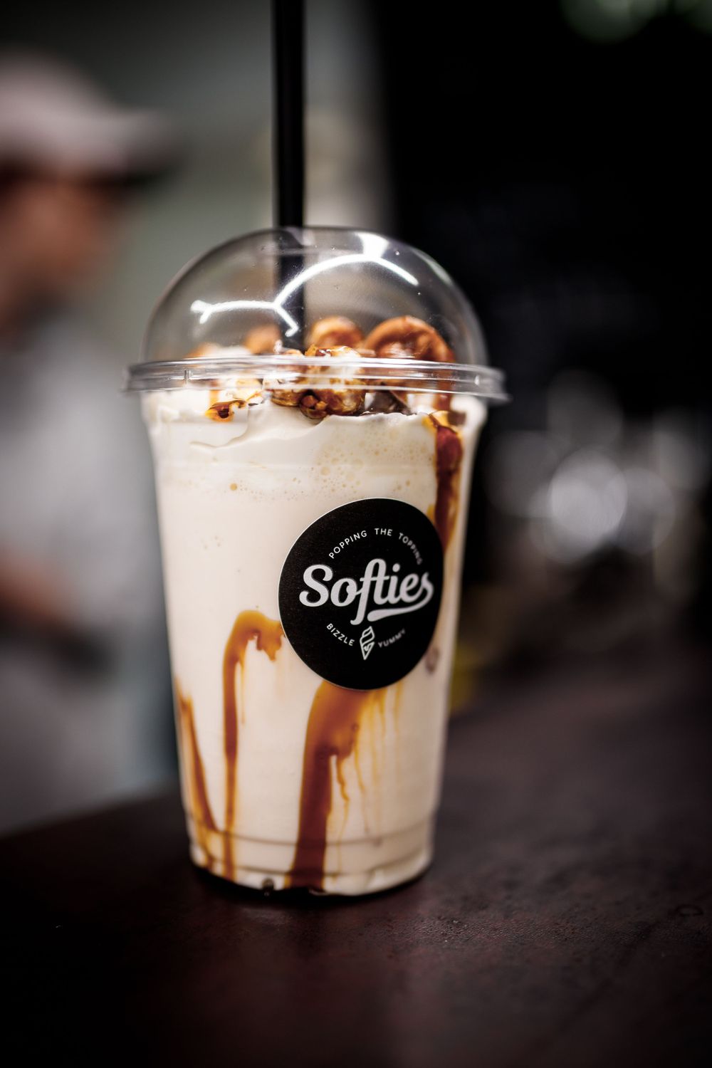 Ein kalter Milchshake mit Schokoladensirup und Schlagsahne in einem Softies-Plastikbecher mit Logo auf einer dunklen Oberfläche.