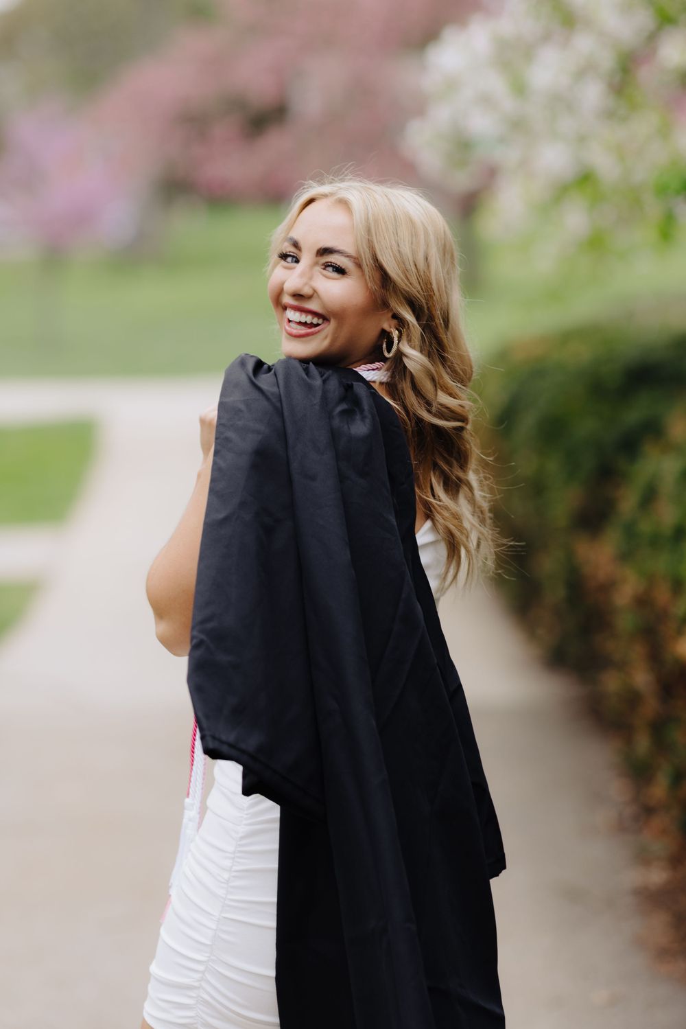 Portrait of graduate in black gown and white dress smiling over shoulder in spring garden setting.