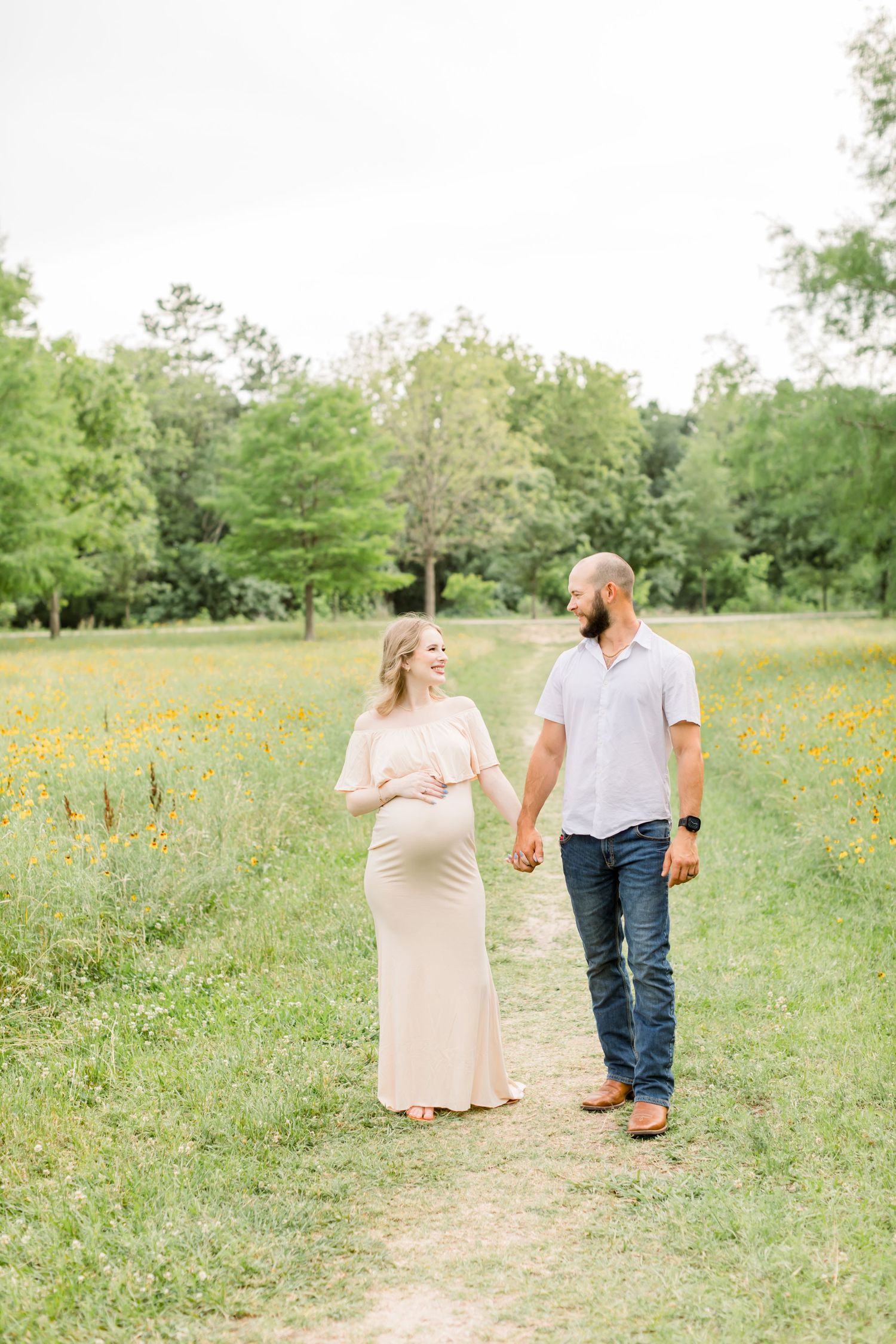 Expectant parents share a casual walk through a meadow filled with yellow wildflowers.