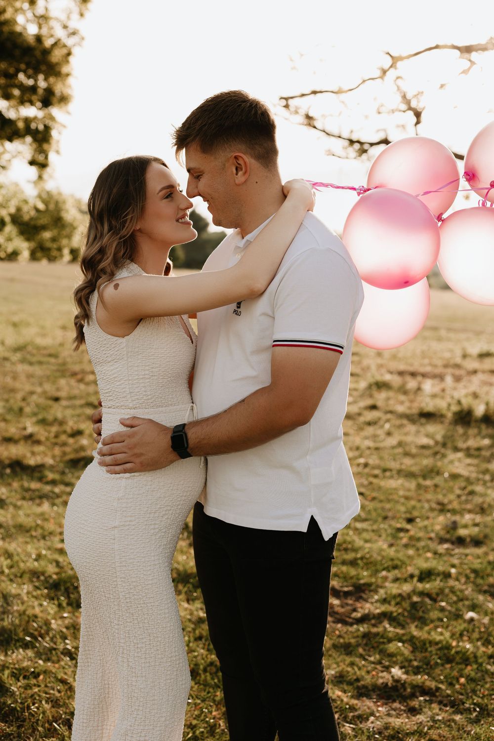 Romantic sunset maternity photos with pink balloons as couple embraces in golden field.