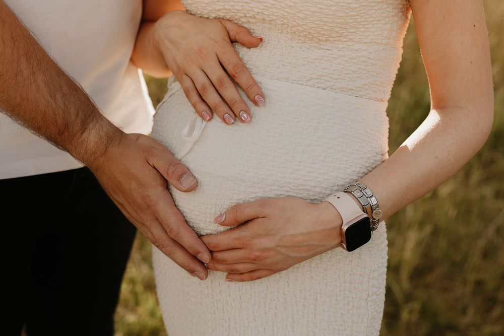 Close up of hands gently cradling a pregnant belly in a white dress against an outdoor background.