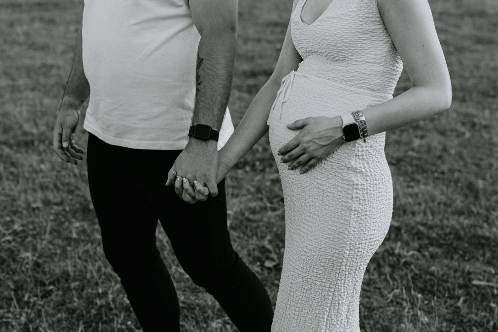 Black and white photo of couple holding hands outdoors with woman in white dress showing pregnant belly.