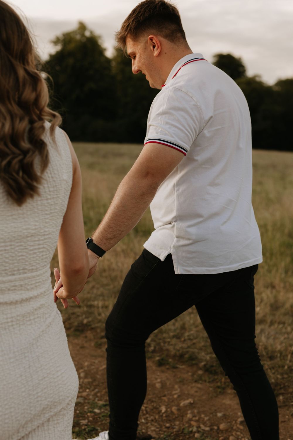 A couple holding hands walking through a field at sunset, with one person wearing a white dress and another in casual attire.