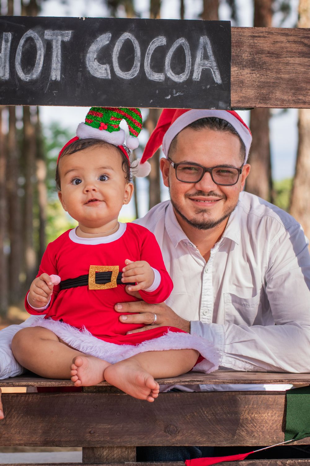 Baby in Santa outfit sits with parent at rustic hot cocoa stand with Christmas decorations.