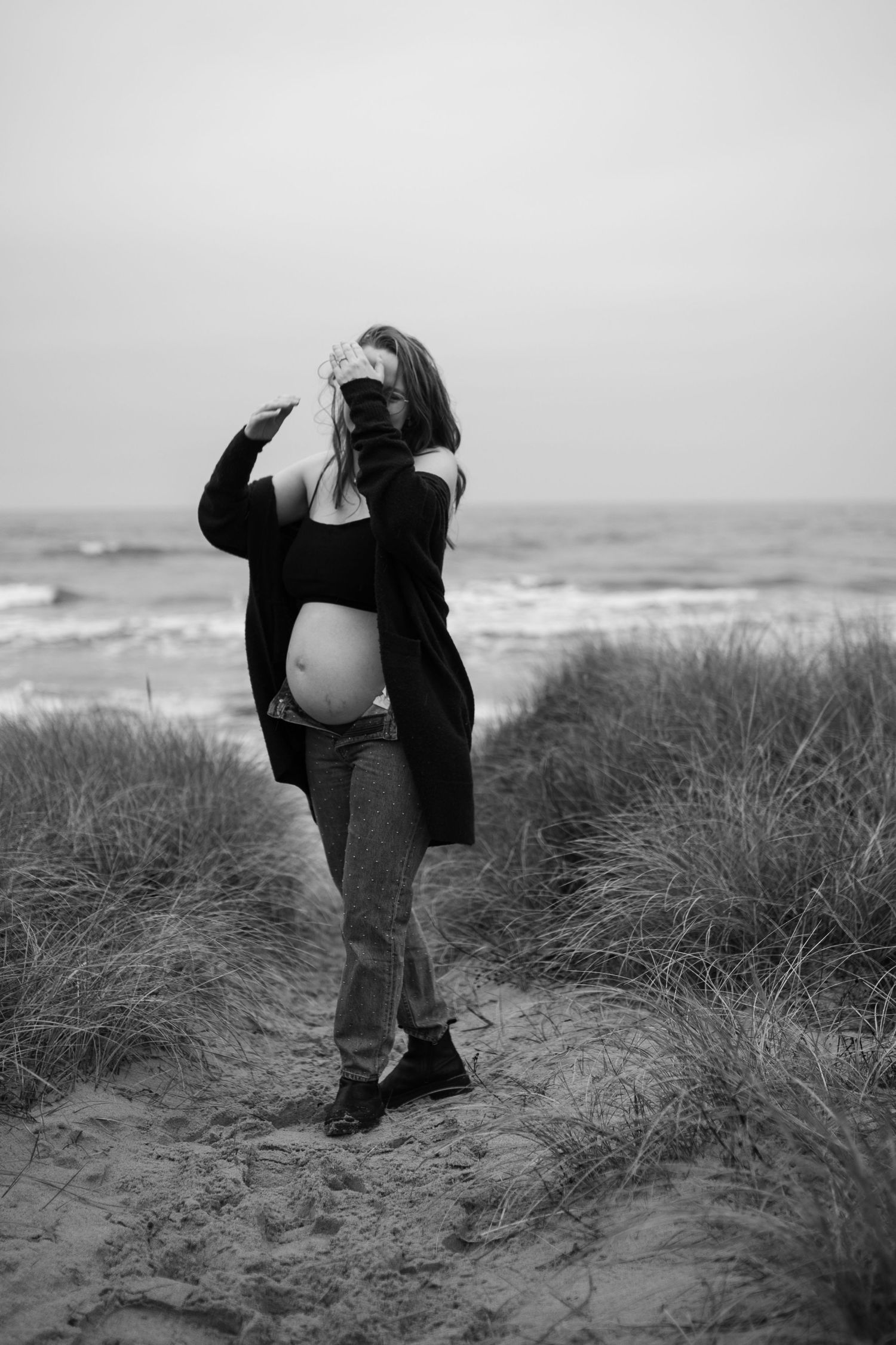 Black and white maternity photo showing someone holding a camera on coastal dunes.