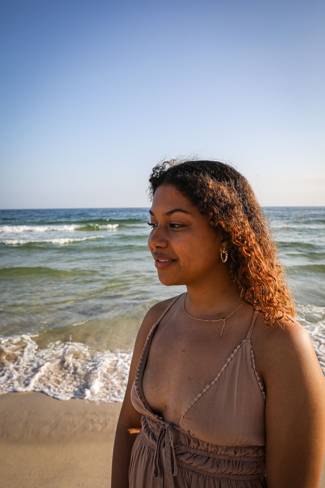 Woman in summer dress stands on beach with ocean waves in background.
