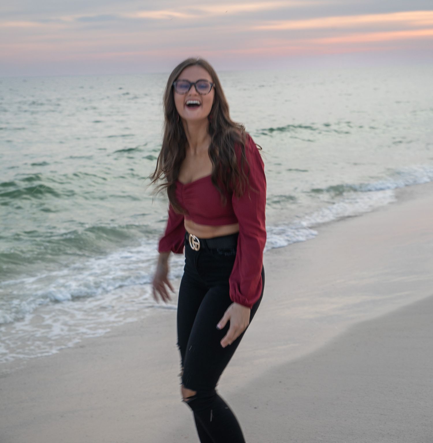 Person in red top and black jeans smiling on beach at sunset.