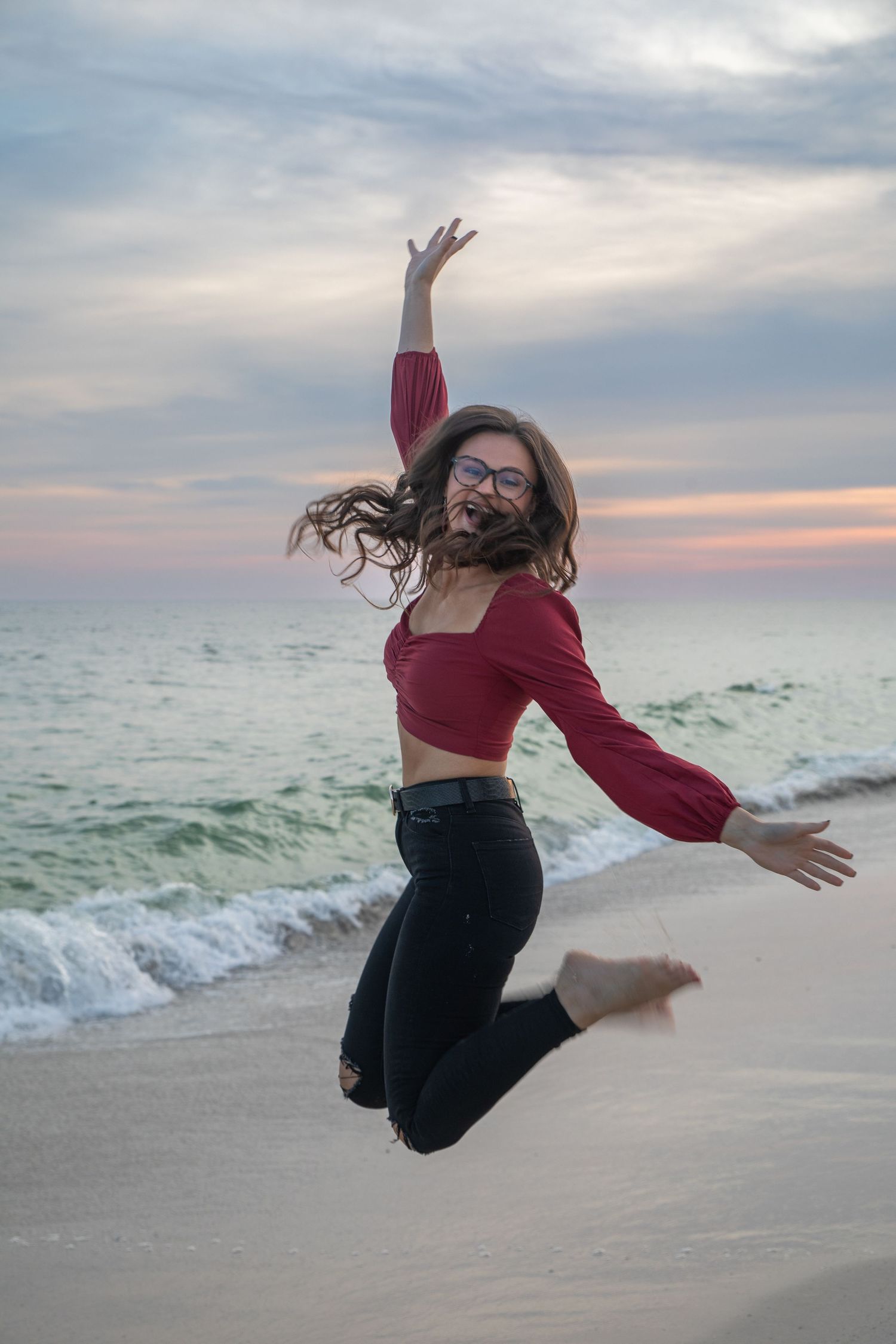 Person in red top and black pants jumps joyfully on beach at sunset with arms outstretched.