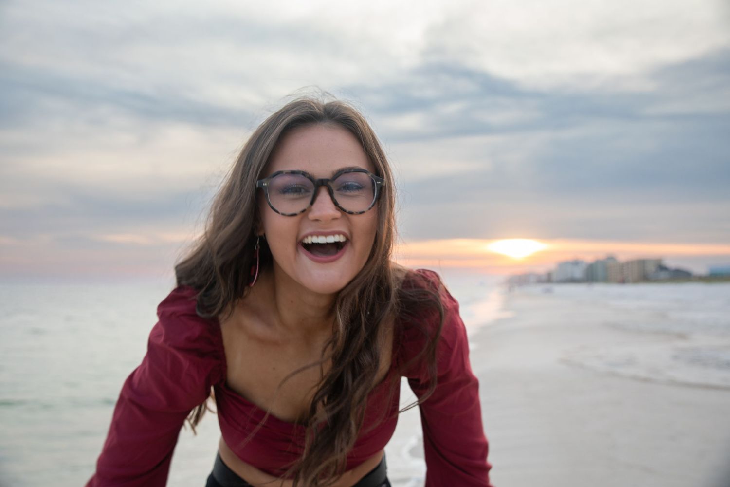 Person wearing glasses and red top laughing joyfully on beach at sunset.