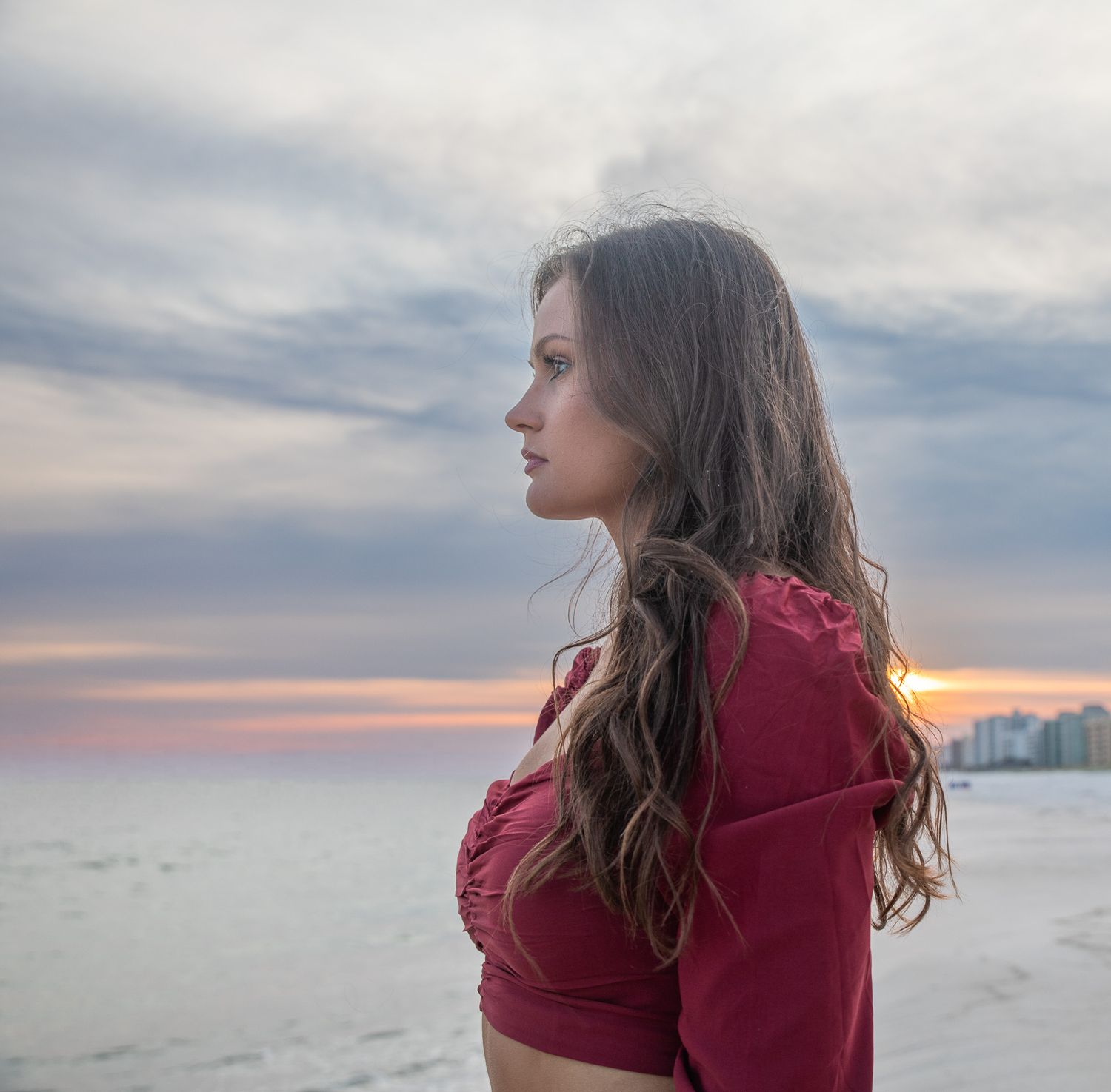 Person in red crop top looking to side with windswept hair on beach.
