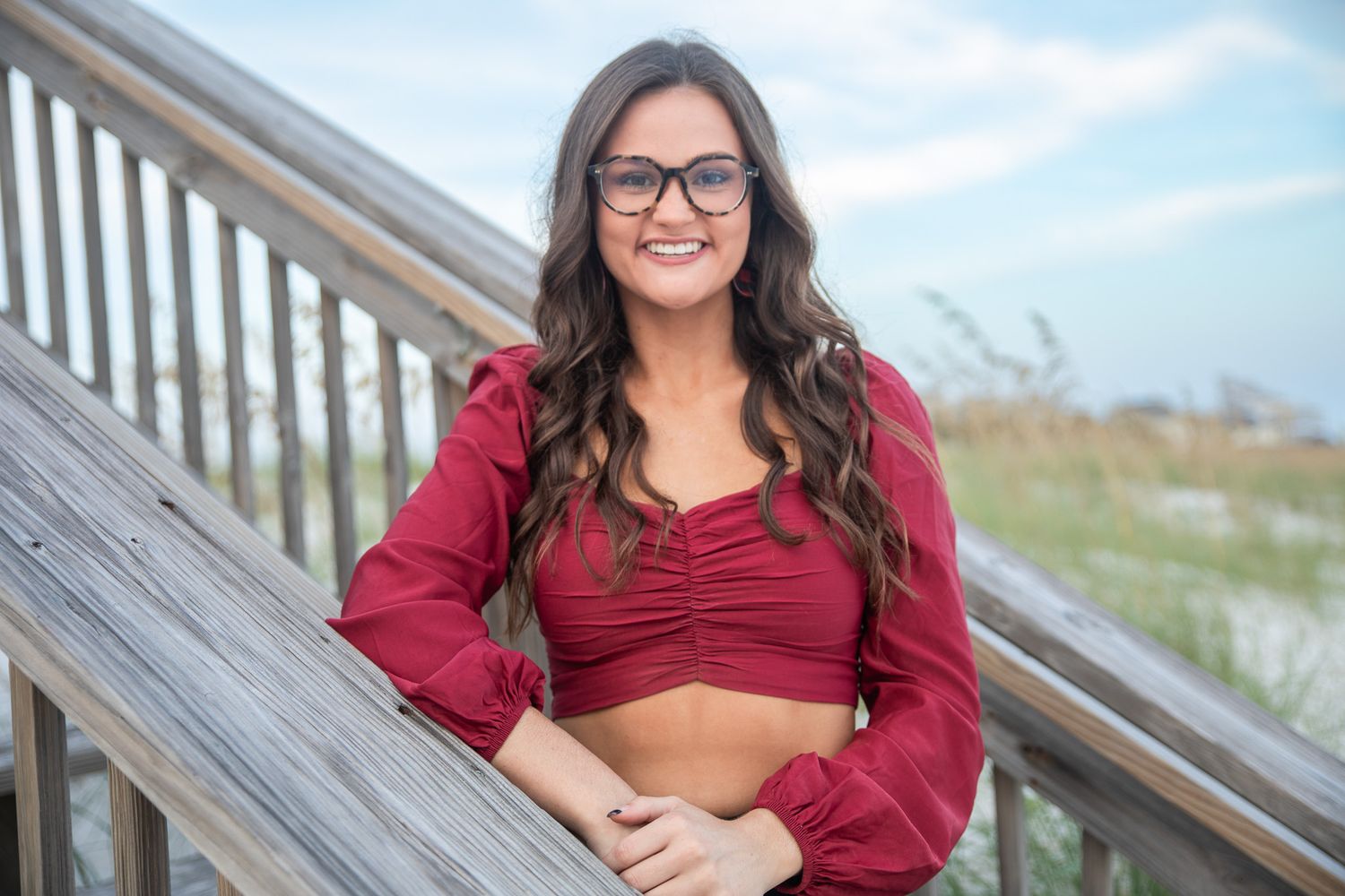 Woman in red crop top with long sleeve balloon sleeves poses on wooden boardwalk at beach setting.