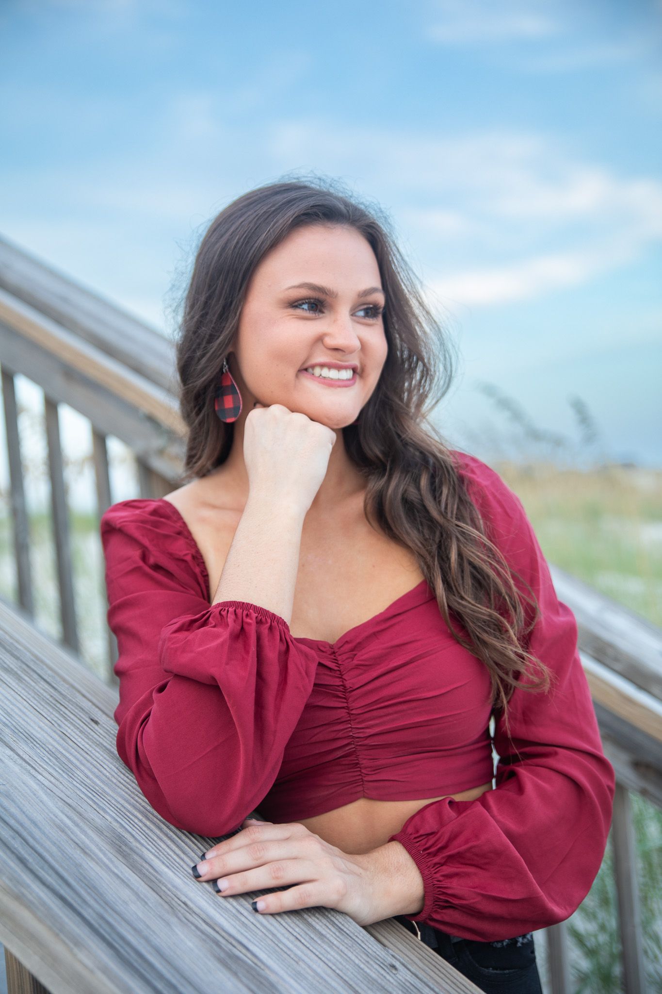 Woman in burgundy crop top poses casually against wooden railing with beach scenery in background.