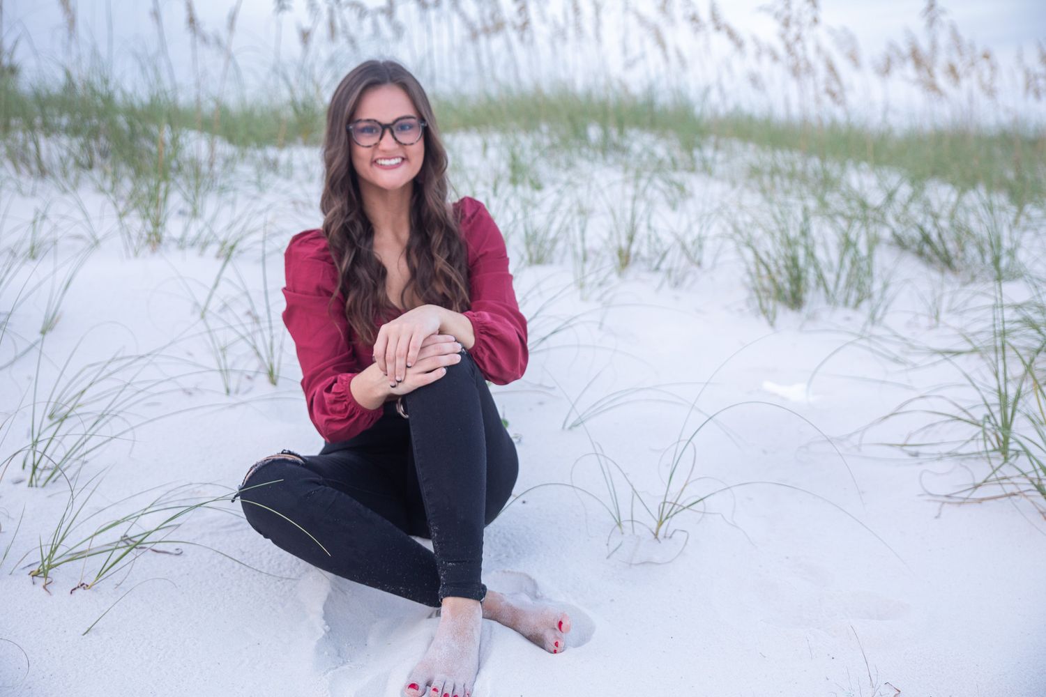 Person wearing glasses and red top sits cross-legged in white sand dunes with beach grass.