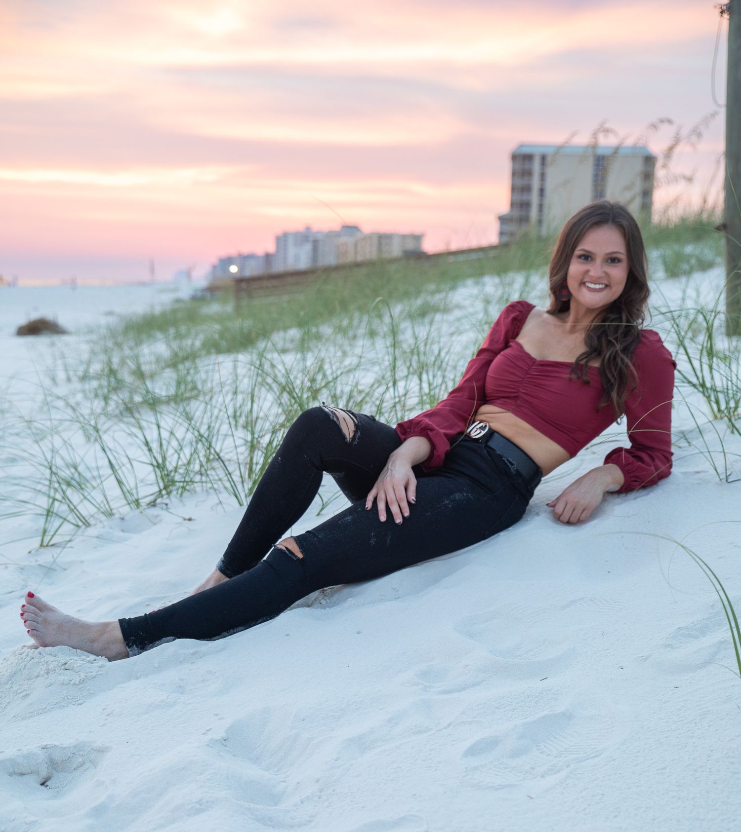 Person in red top and black pants reclines on white sand beach at sunset.