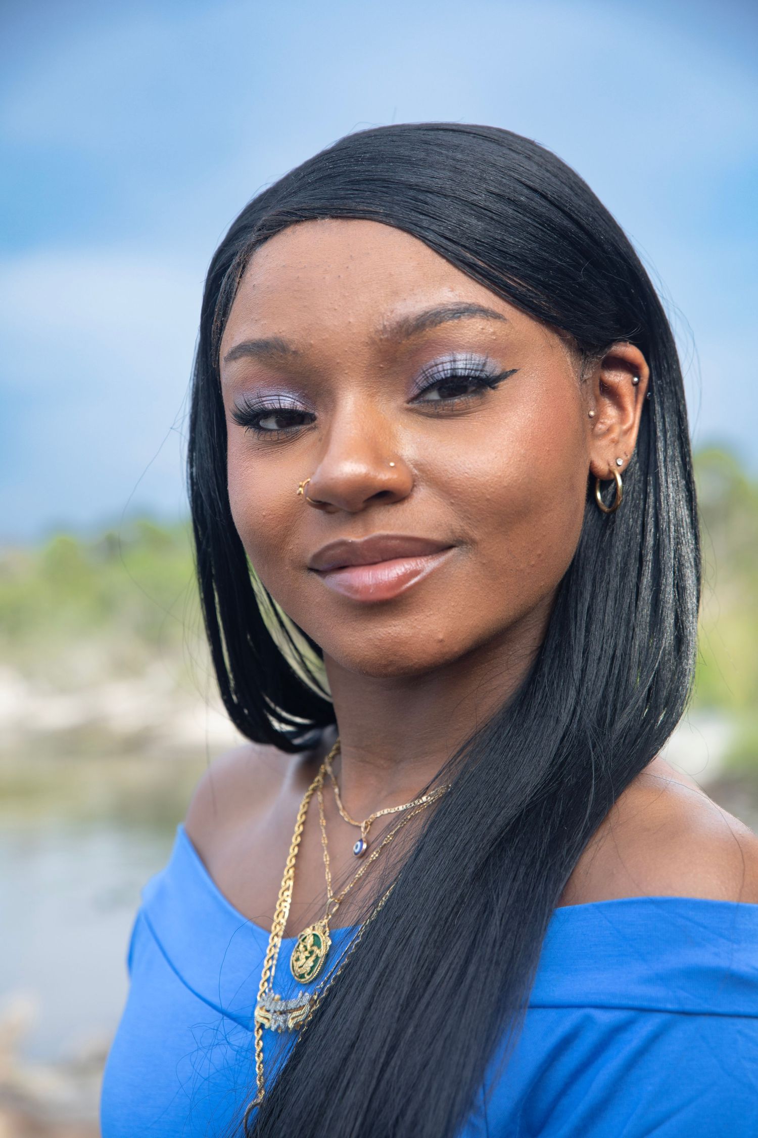 Portrait of someone wearing a royal blue off-shoulder top with layered gold necklaces against a blurred outdoor background.