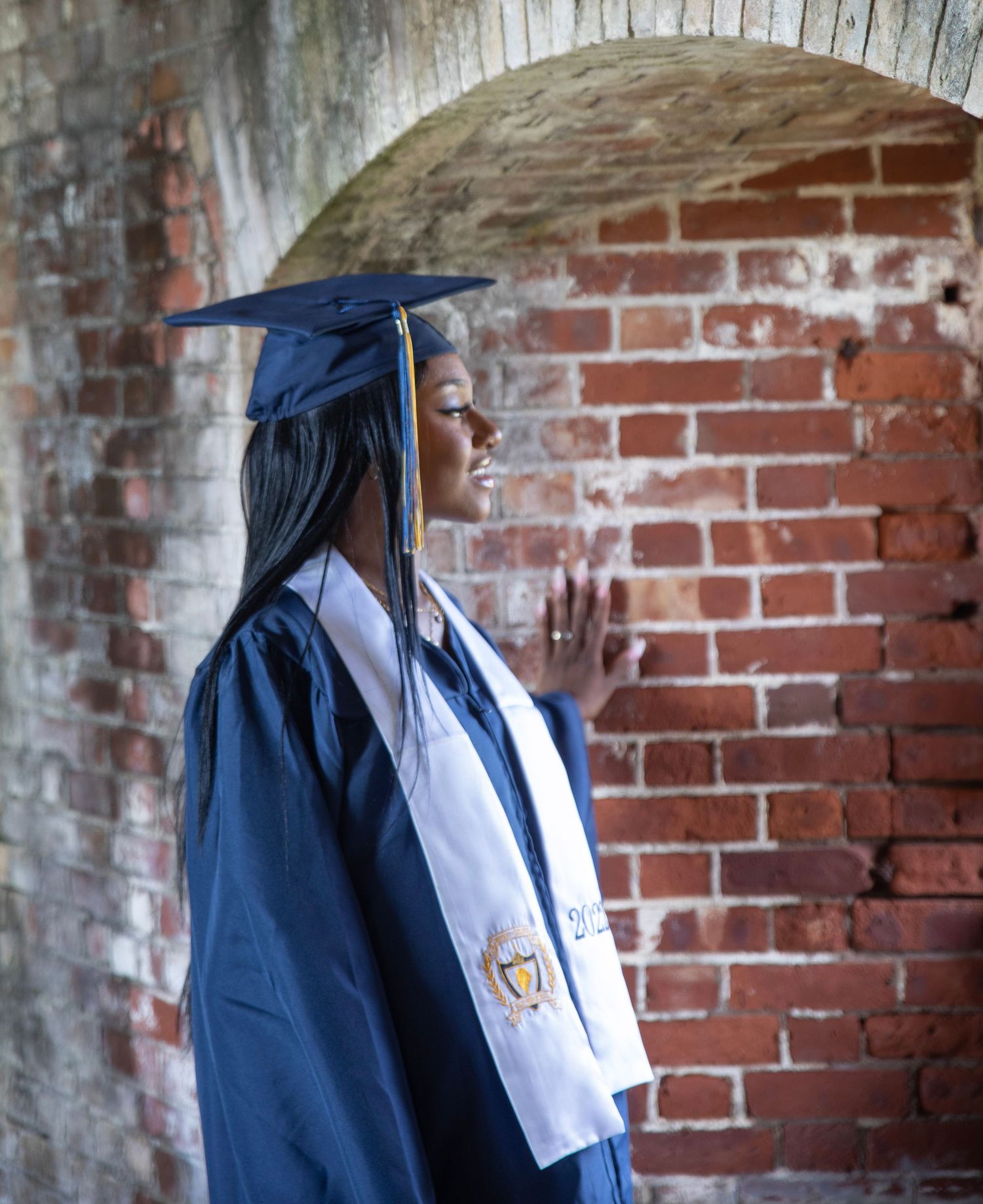 Graduate in navy blue cap and gown photographed in profile against weathered brick archway.