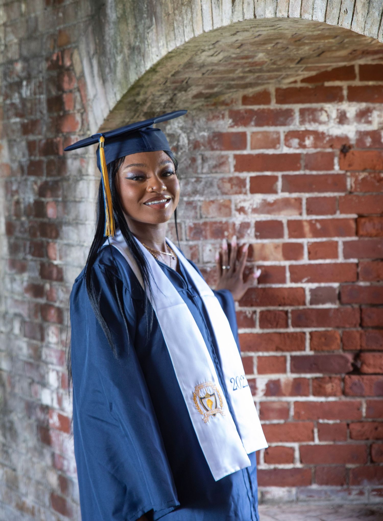 Graduate in blue academic regalia smiling next to brick wall with historic arch.