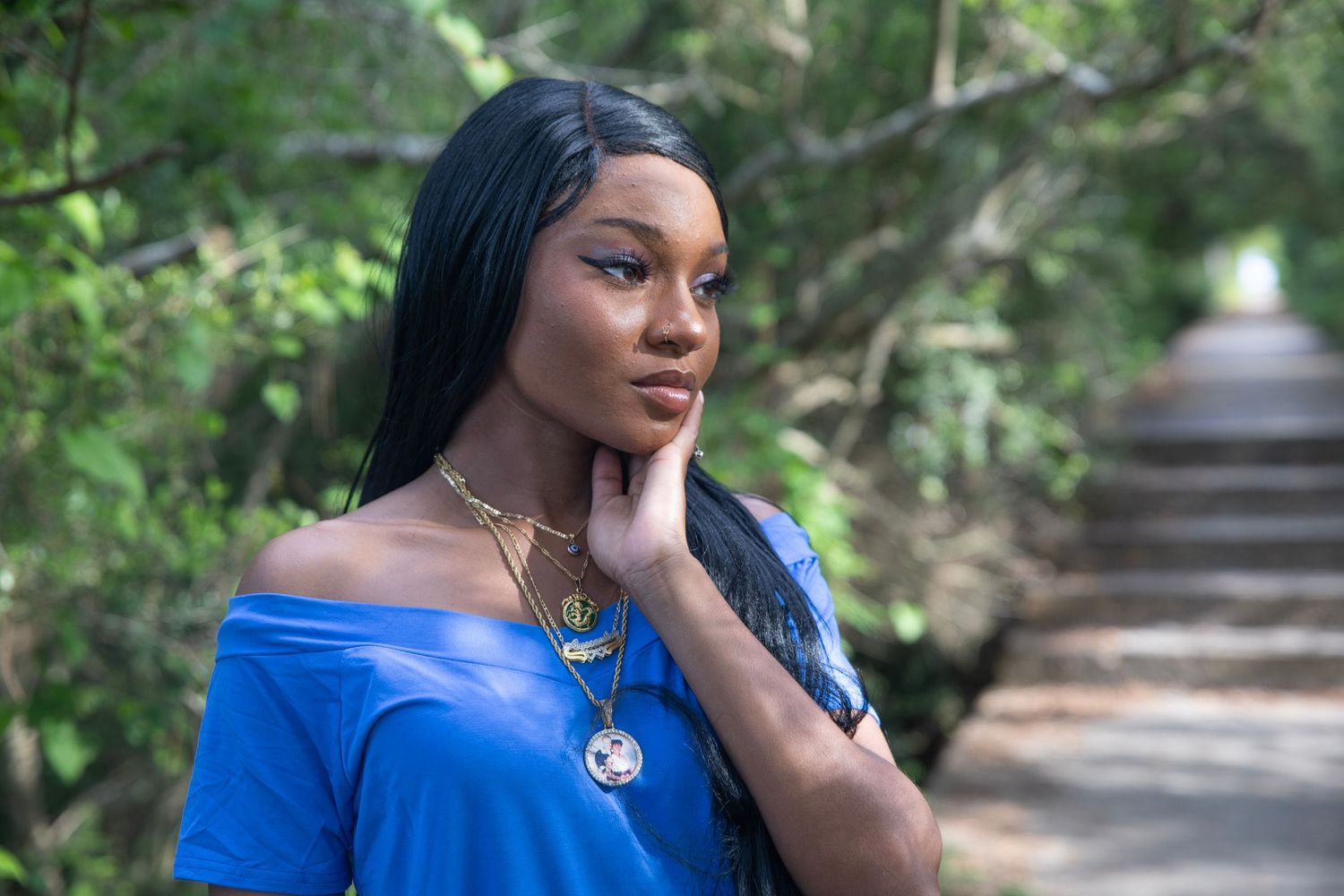 Portrait of someone in blue off-shoulder top with gold jewelry against blurred nature background.