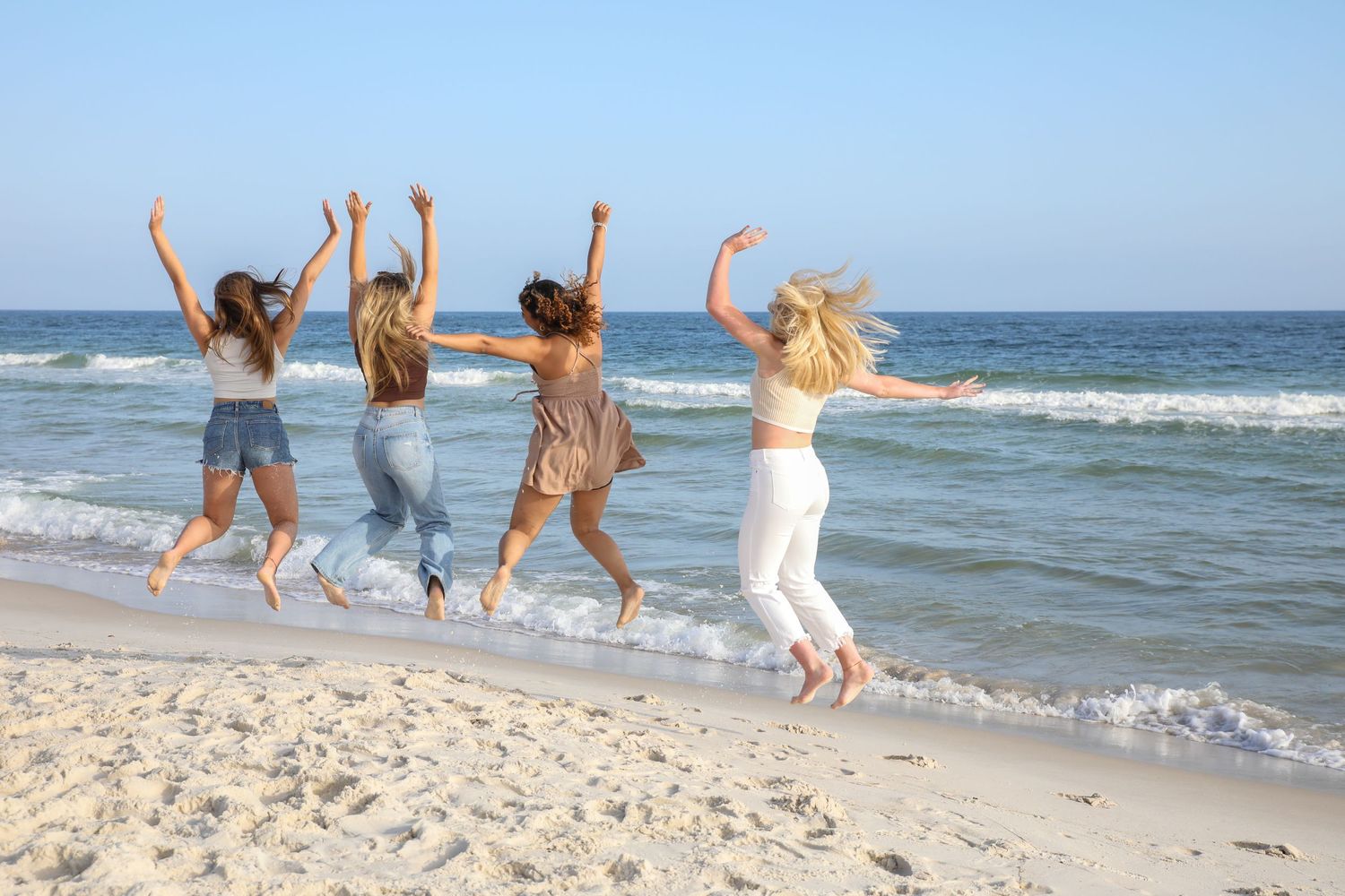 Four friends jump joyfully on a sunny beach with ocean waves in the background.